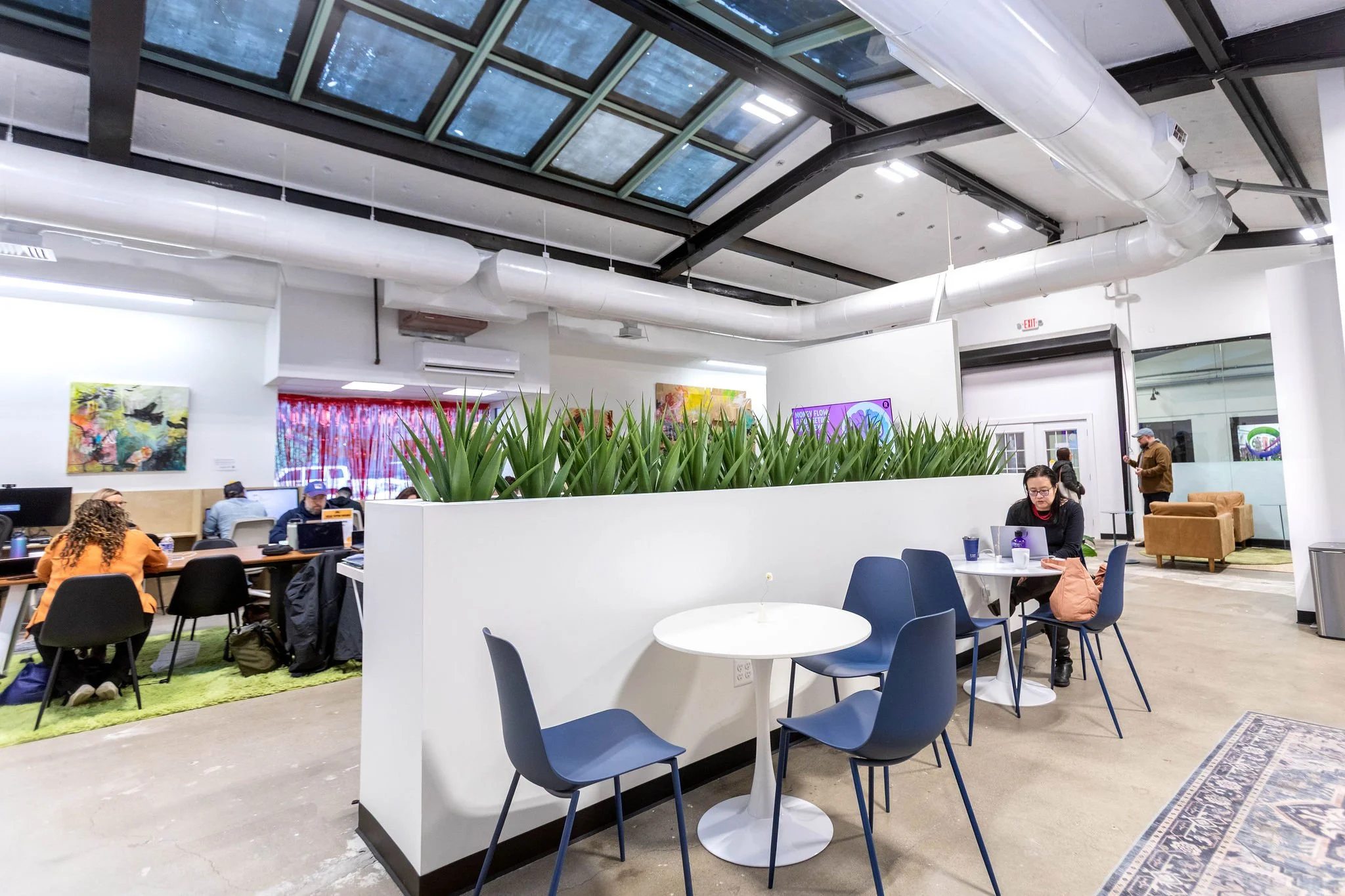 People working and studying in a modern, open-concept office or coworking space with large skylights, colorful artwork on the walls, a white partition with greenery, and seating areas with chairs and tables.