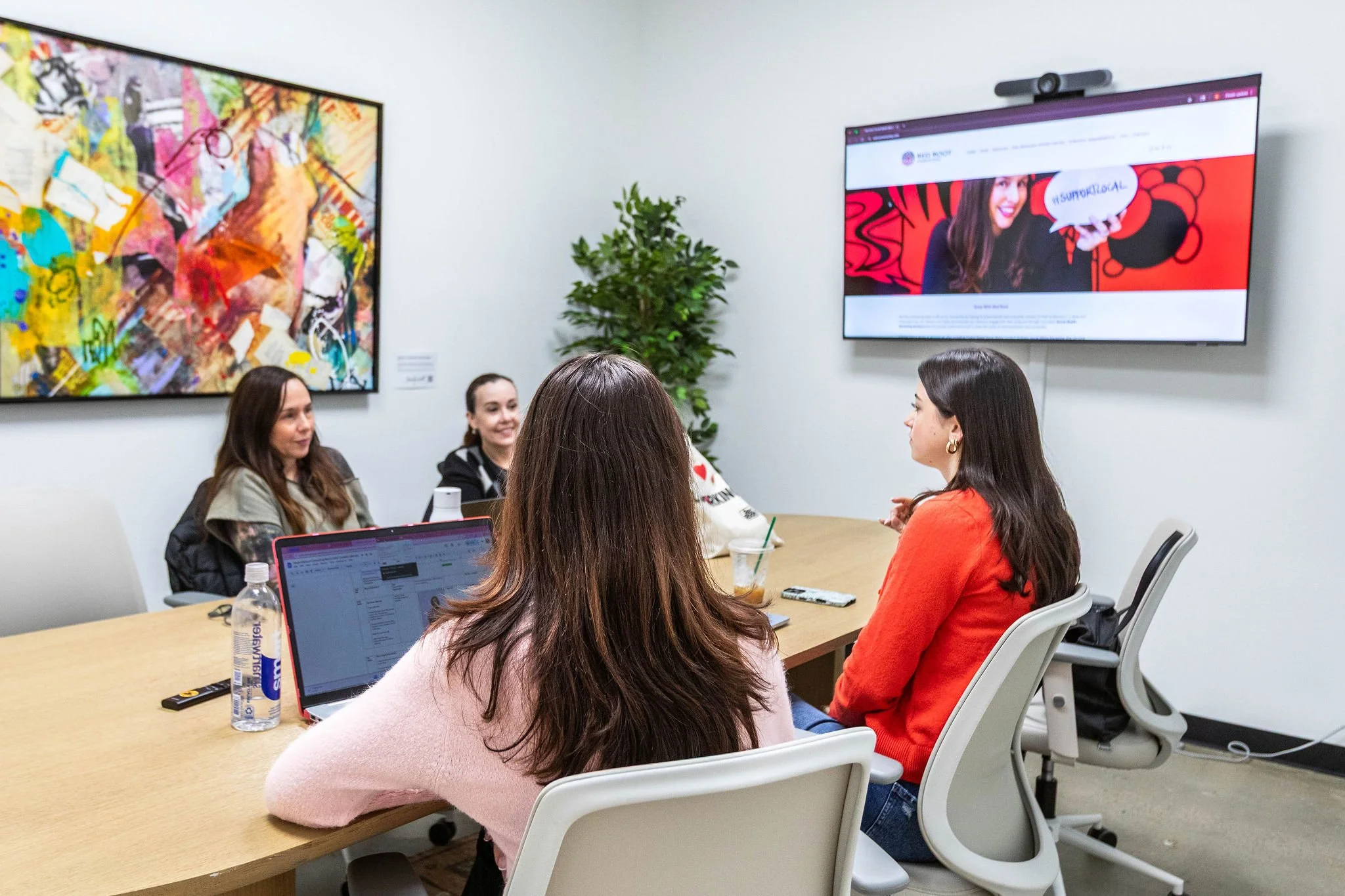 A group of women sitting around a conference table in a meeting room, with a large screen on the wall displaying a website, and a piece of colorful abstract art on the adjacent wall.
