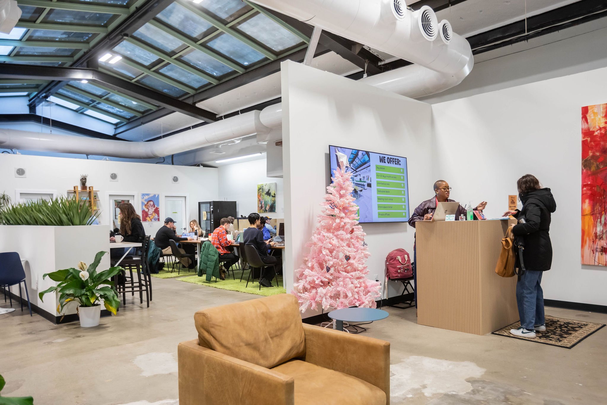 Interior of a modern shared workspace or cafe with people working on laptops, a pink Christmas tree near the reception desk, and colorful artwork on the walls.