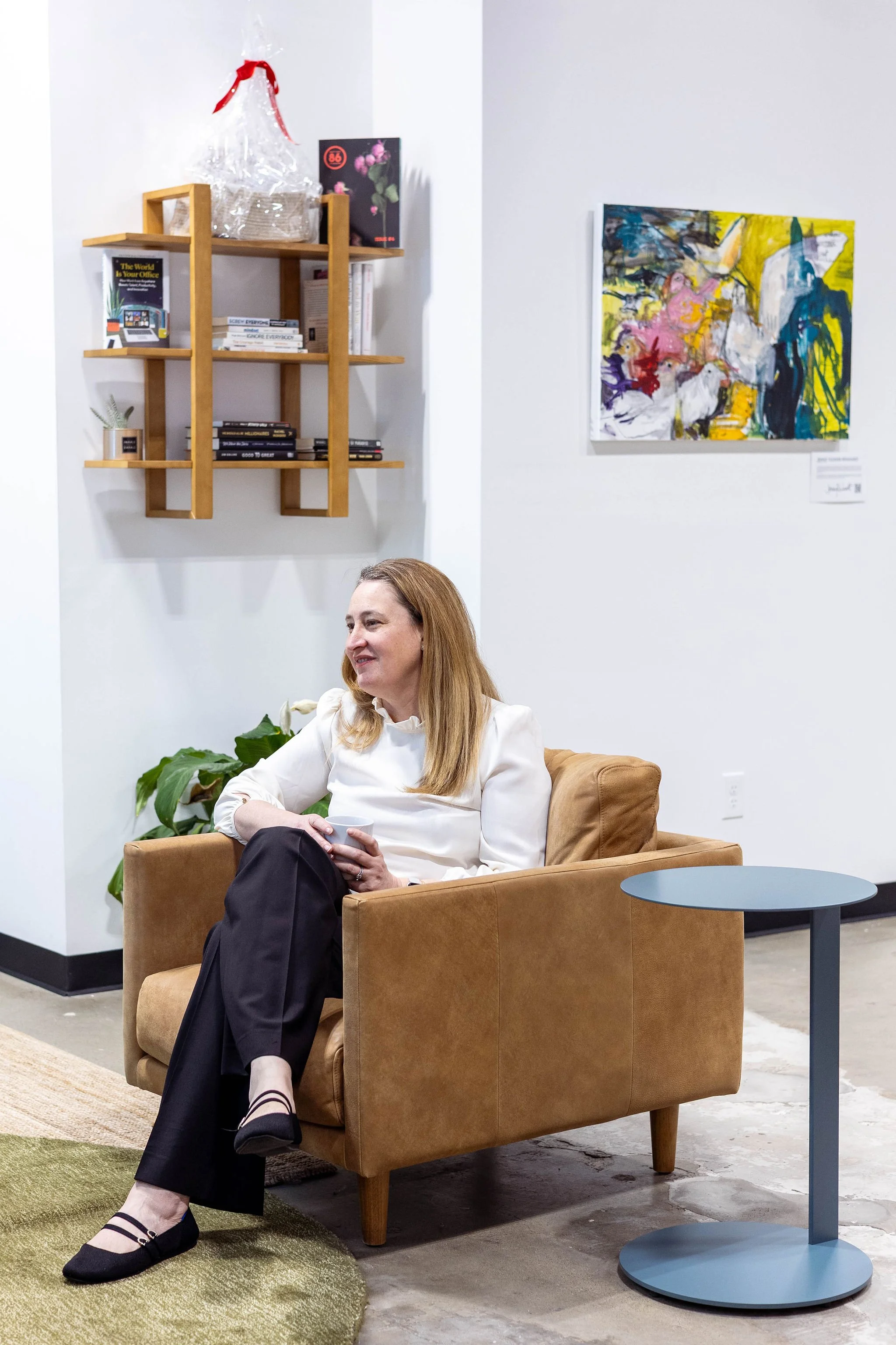 A woman with blonde hair sitting in a tan armchair, holding a mug, in an art gallery or similar setting, with colorful abstract paintings and wooden shelves with books on the wall behind her.