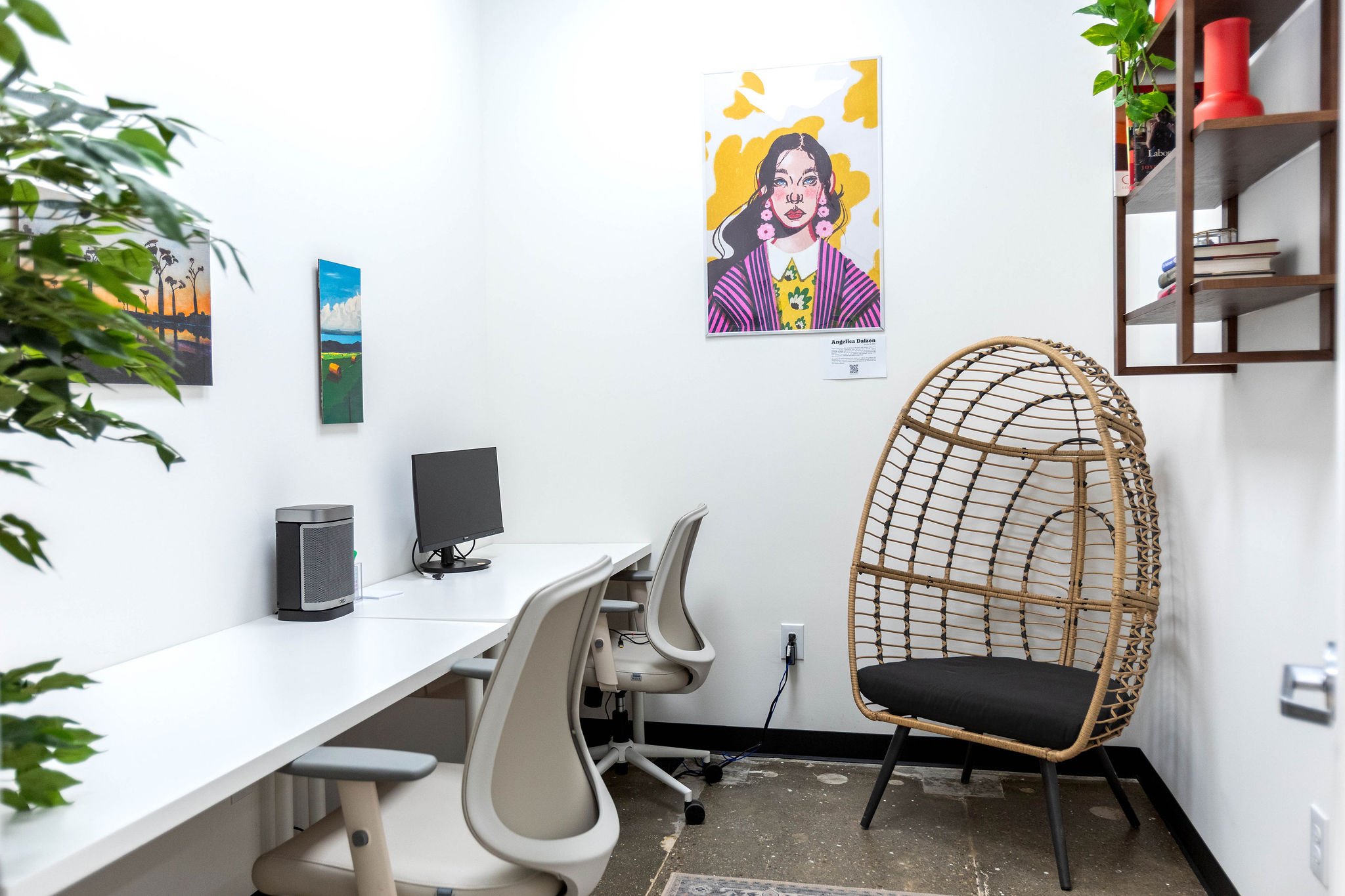 An office with two white desks, two white chairs, a small computer, and a speaker. There are colorful paintings on the wall, including a portrait of a woman with dark hair, earrings, and a colorful outfit. A rattan chair with a black cushion is besid