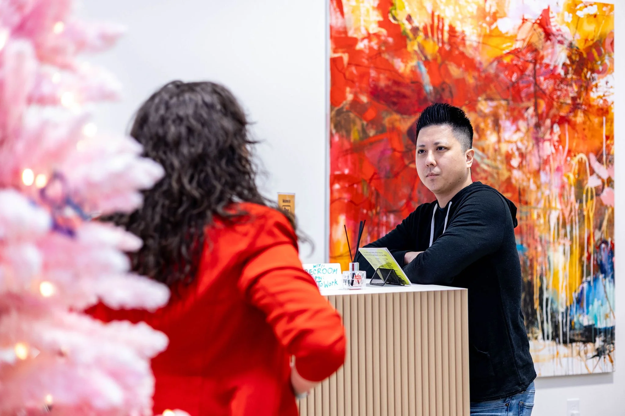 A man standing at a reception desk in an art gallery, talking to a woman, with a large, colorful abstract painting in the background and a pink Christmas tree in the foreground.