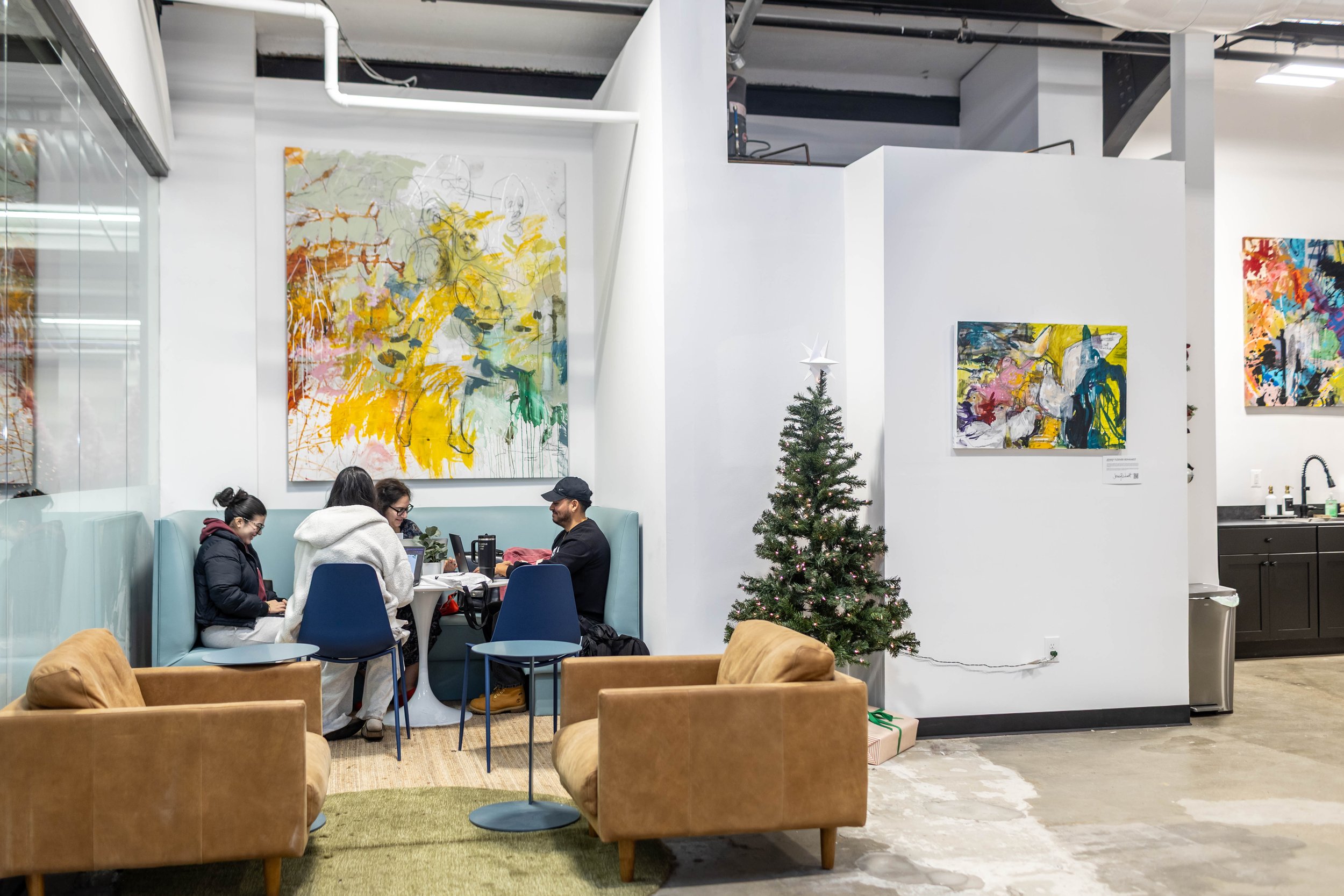 People sitting at a table in a café with decorated Christmas tree, colorful abstract paintings on white walls, and modern furniture.
