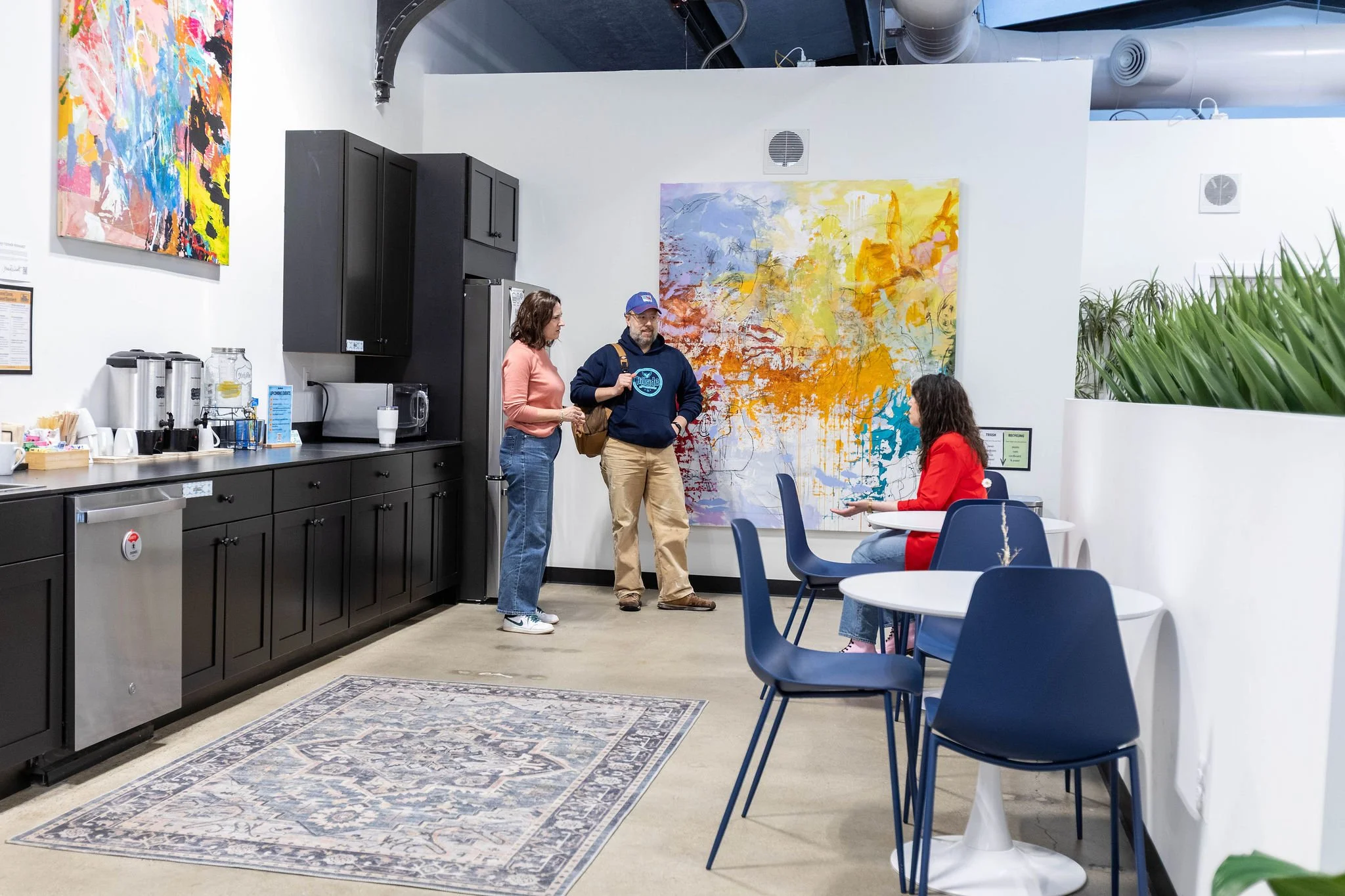 Interior of a modern café or lounge area with two people standing and talking, and one seated woman. The space features black cabinets, a colorful abstract wall and paintings, a rug, and a coffee station with drinks on the counter.