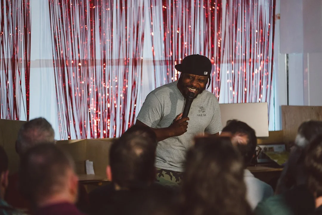 Man smiling and speaking into microphone during a presentation or event with an audience. Red curtain background.
