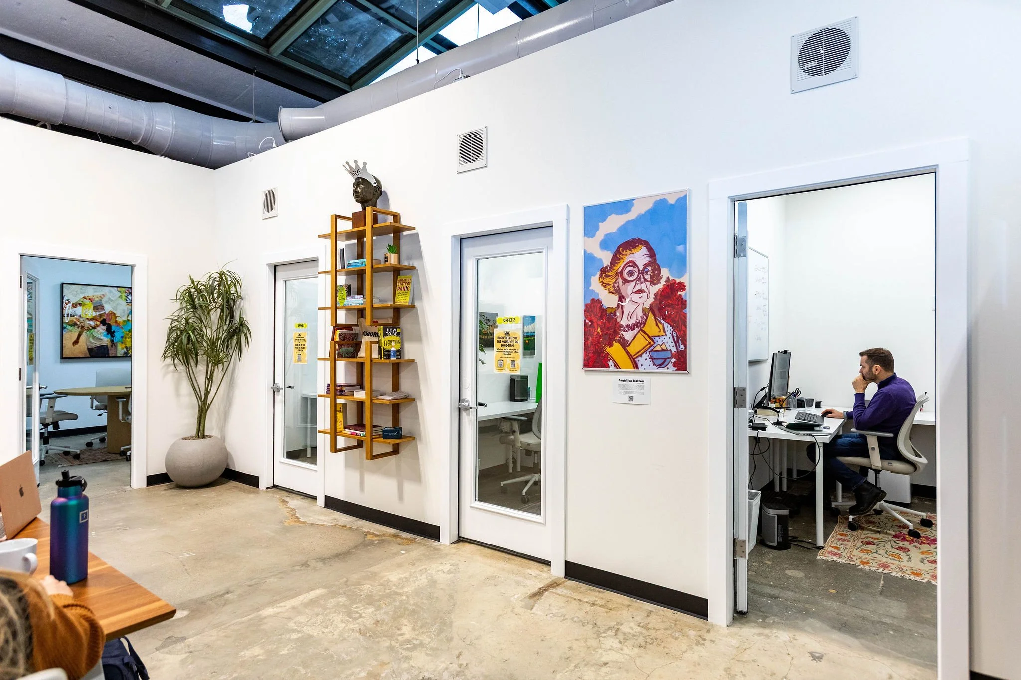 View of an office space with multiple rooms, a bookshelf on the wall, artwork including a colorful portrait of an elderly woman, and a man working at a desk inside a room to the right.