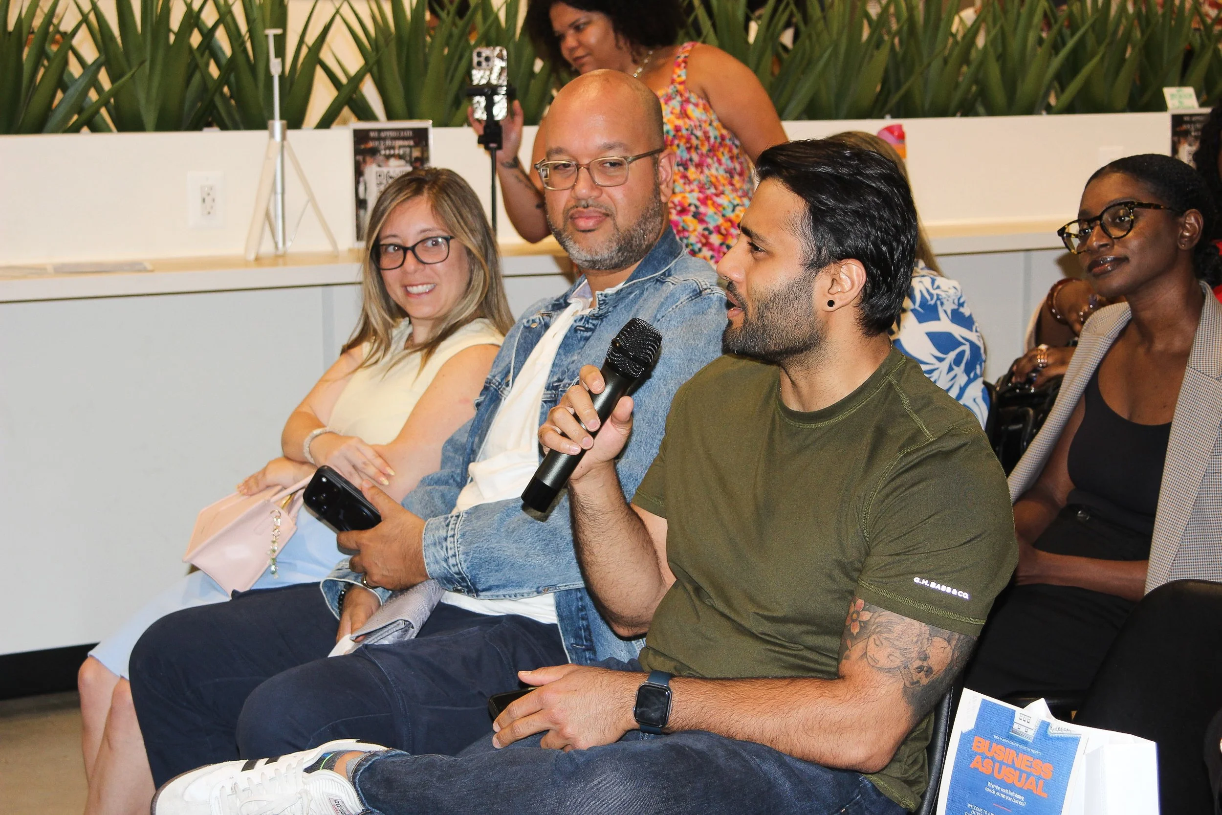 A group of diverse people sitting in a row at a conference or seminar. One man is holding a microphone and talking, others are listening and observing. There are books and plants behind them.