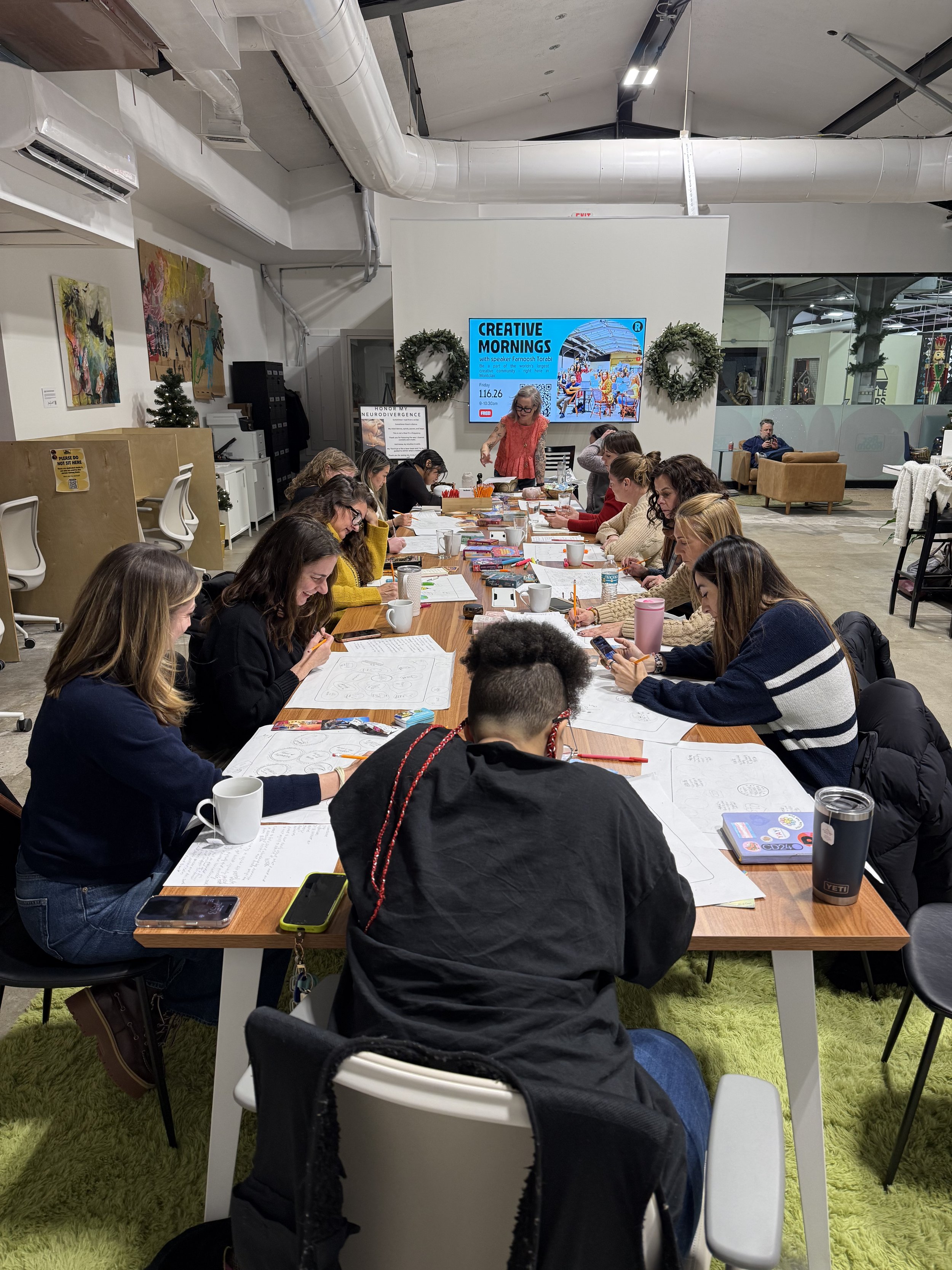 A group of people sitting around a long table participating in a workshop or meeting in a modern, well-lit room. There are papers, notebooks, and drinks on the table, and a woman standing at the head of the table speaking. The room is decorated with Christmas wreaths, artworks on the wall, and a large digital screen displaying information about 