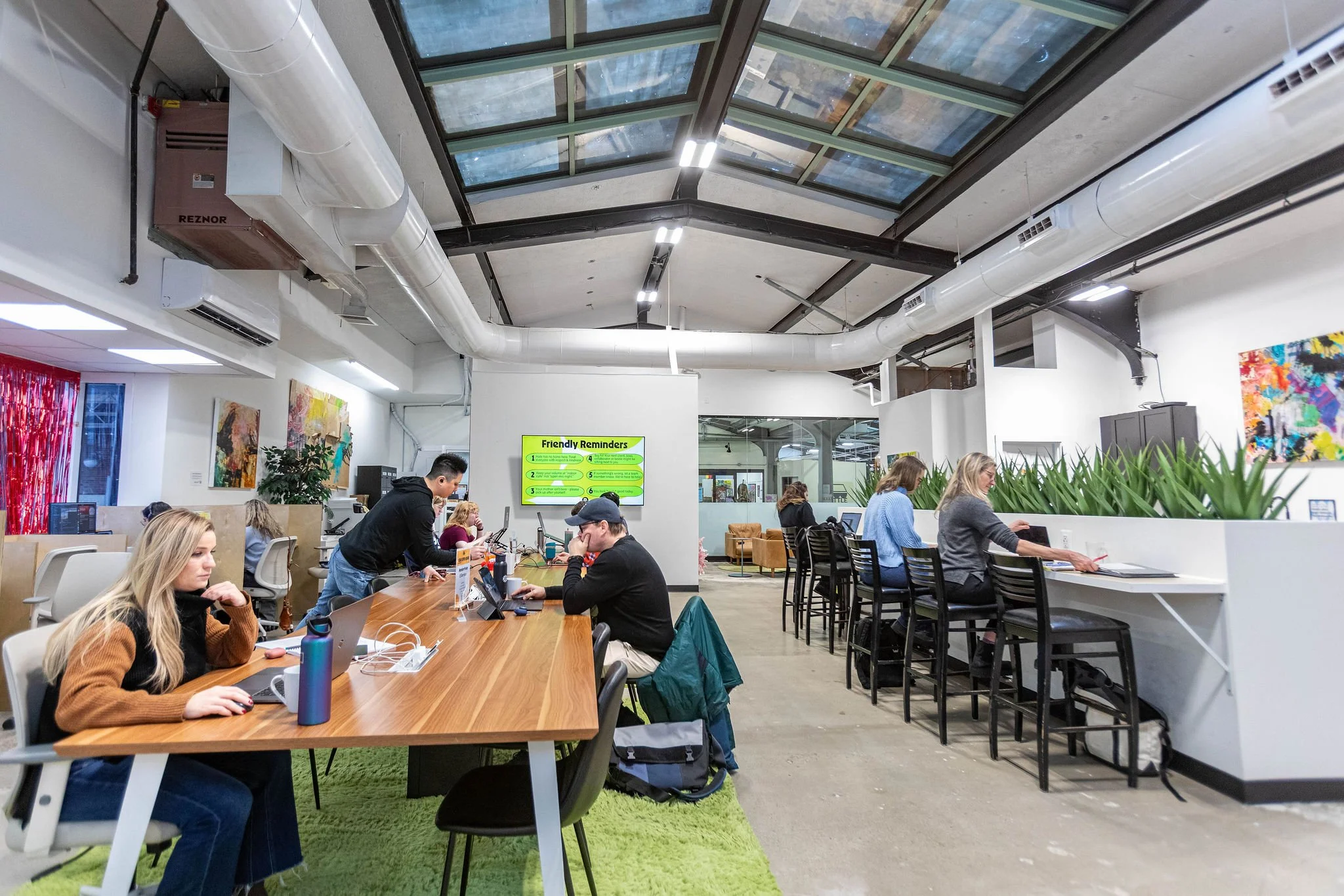 People working on laptops and tablets in a modern coworking space with a high ceiling, skylights, artwork on white walls, and a green rug.