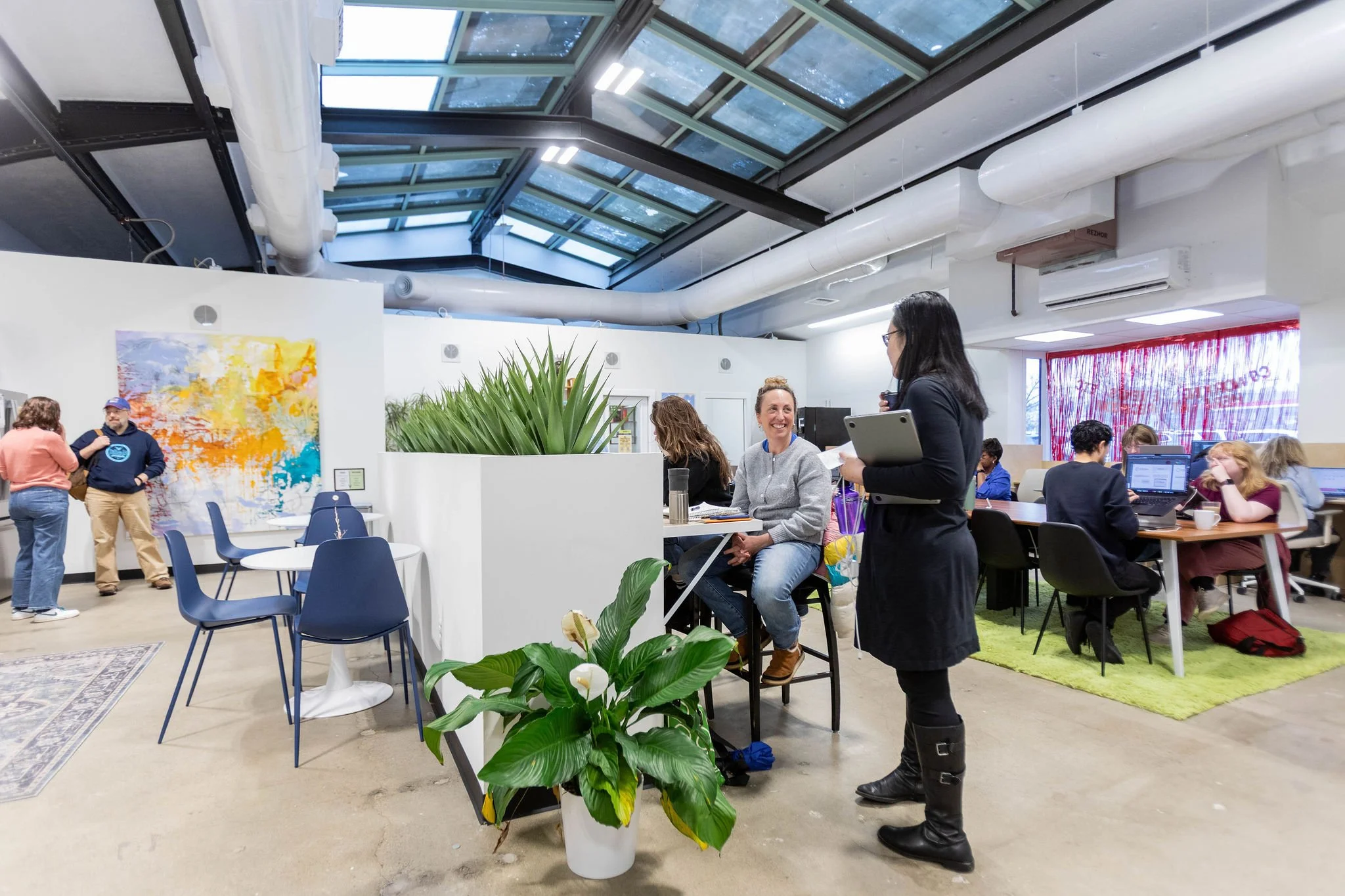 People working and socializing in a modern, open-plan office space with large skylight windows and colorful decor.