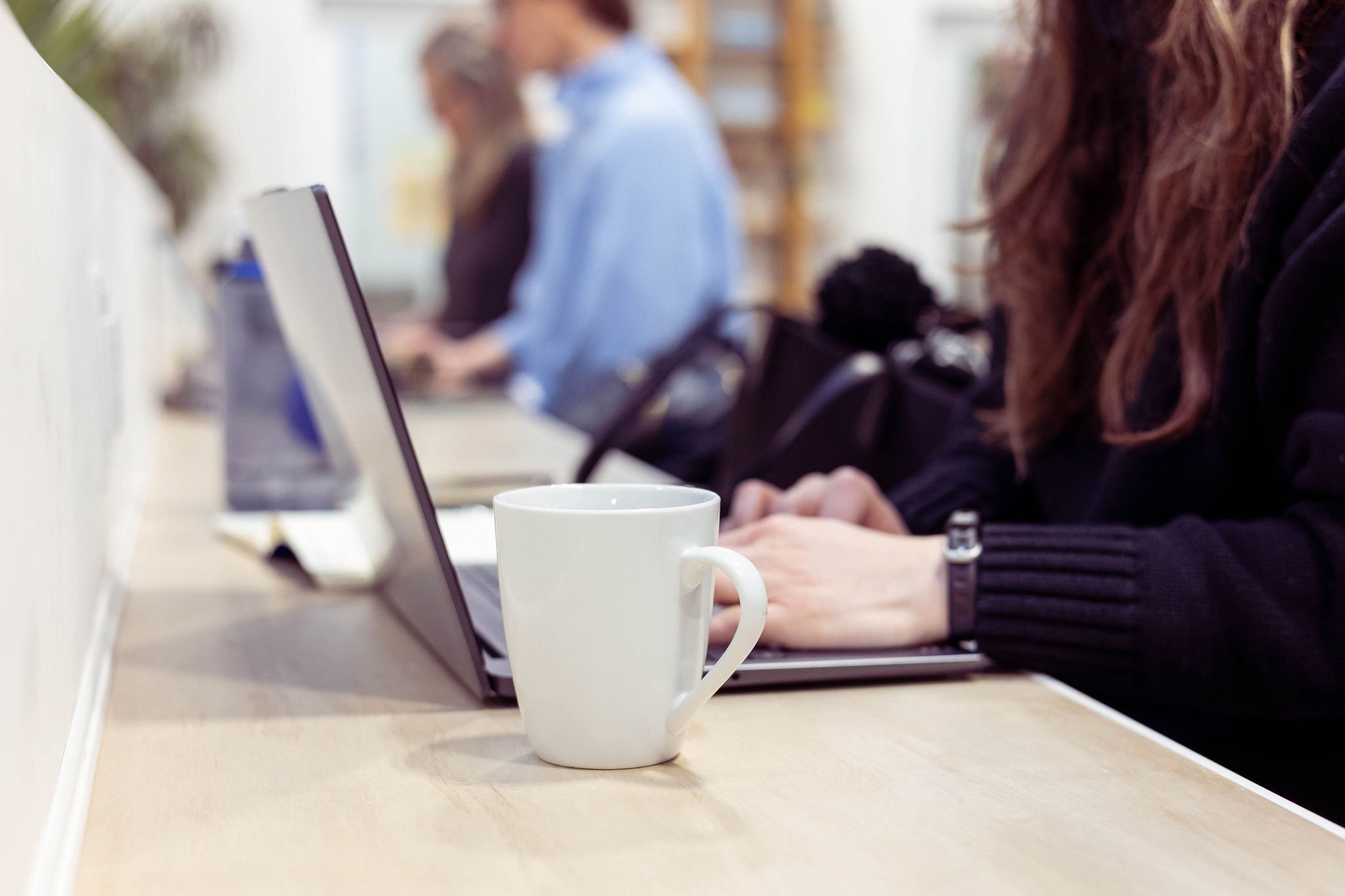 A white coffee mug and a laptop on a light-colored table in a cafe or workspace, with people working on laptops in the background.