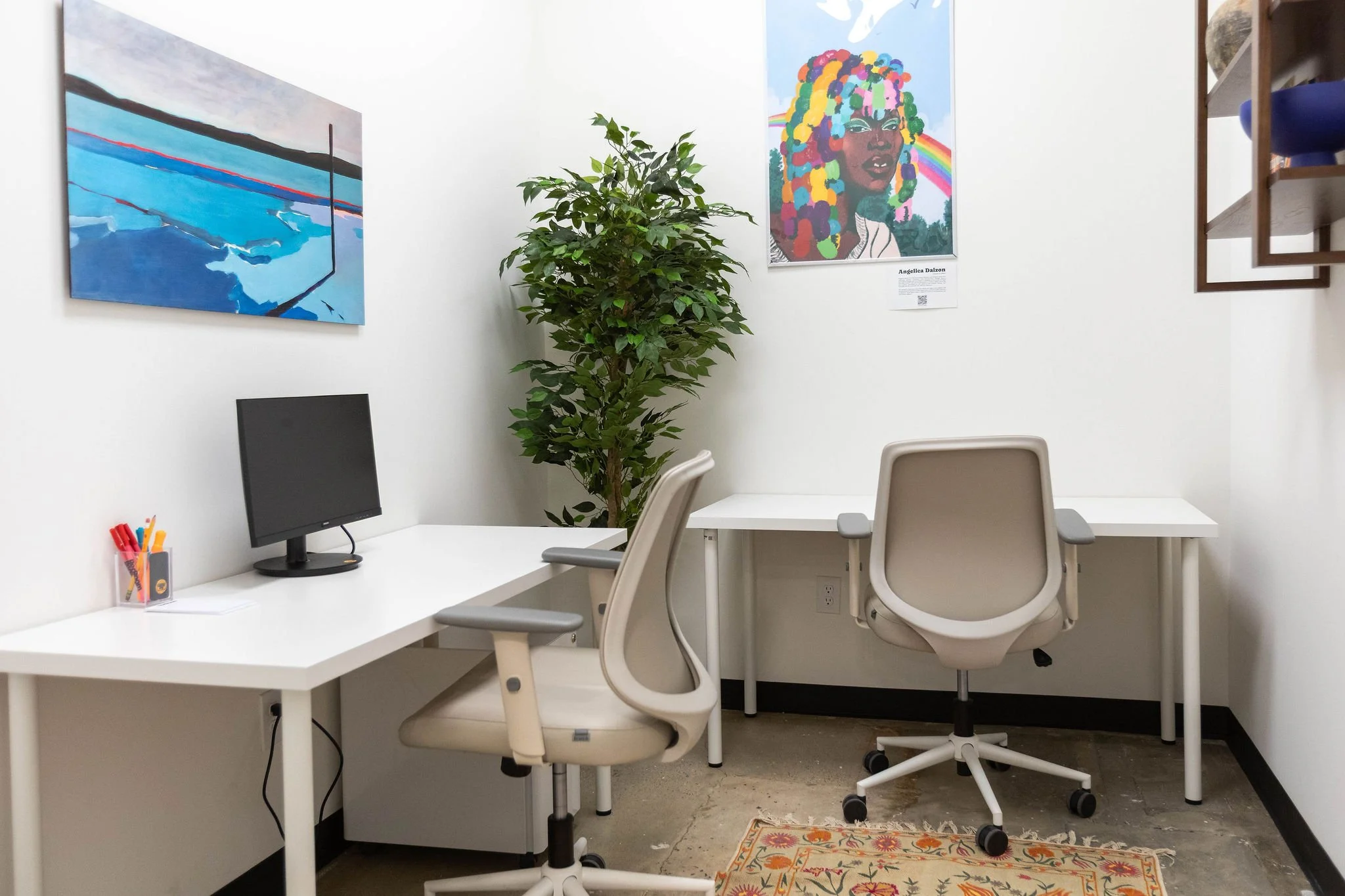 An office with two white desks, two beige chairs, a computer monitor, a cup of colorful pens, wall art, a large potted plant, a patterned rug, and shelves with decorative items on the right.