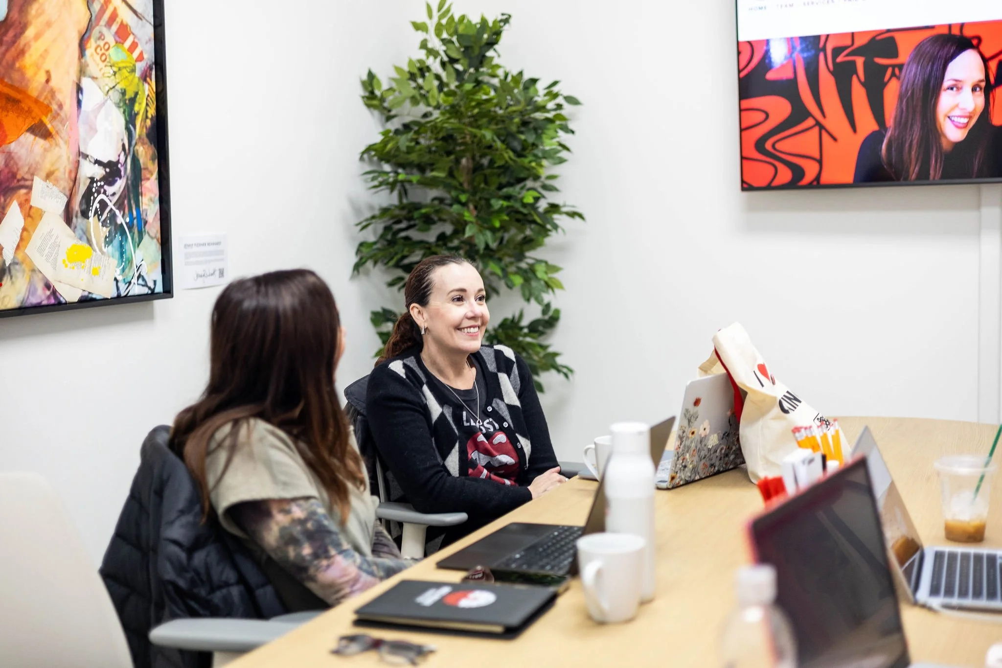Two women sitting at a conference table, engaged in a discussion, with a woman smiling, a laptop, notebooks, coffee mugs, and a bag on the table; artwork on the wall, a television, and a potted plant in the background.