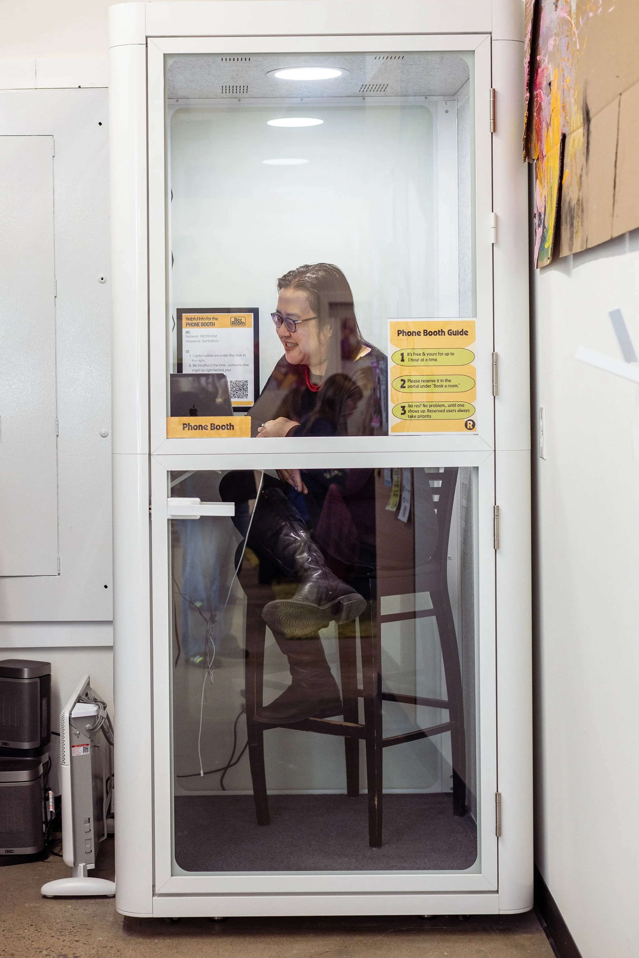 A woman sitting inside a phone booth, seen through the glass, with signs on the booth door.