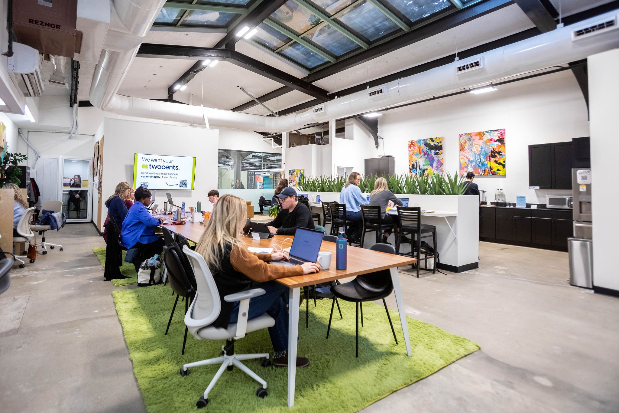 People working at desks in a modern, shared office space with contemporary art on the walls and a green rug on the concrete floor.
