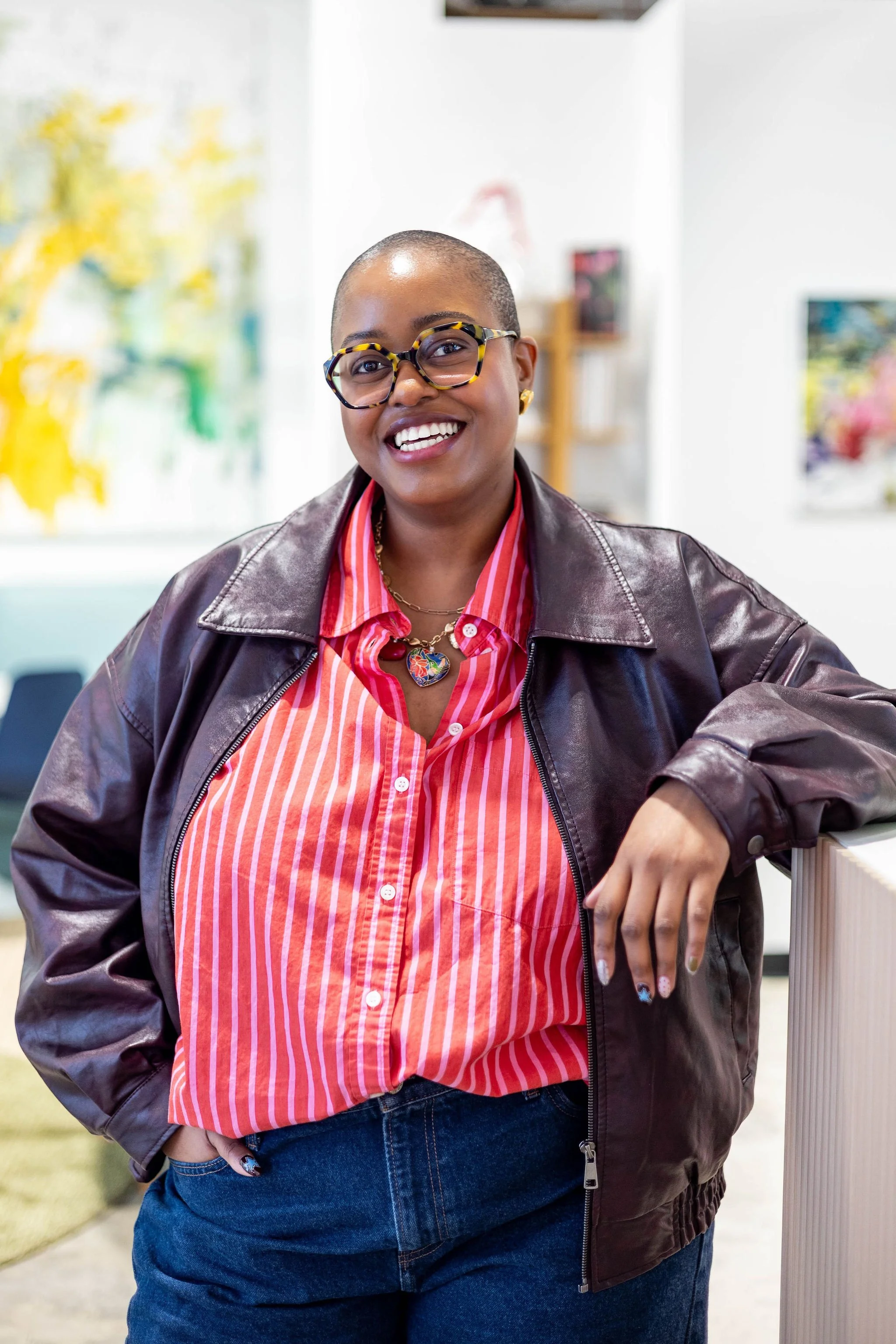 A smiling woman with glasses, short hair, wearing a leather jacket over a red and white striped shirt, accessorized with a colorful heart-shaped necklace.