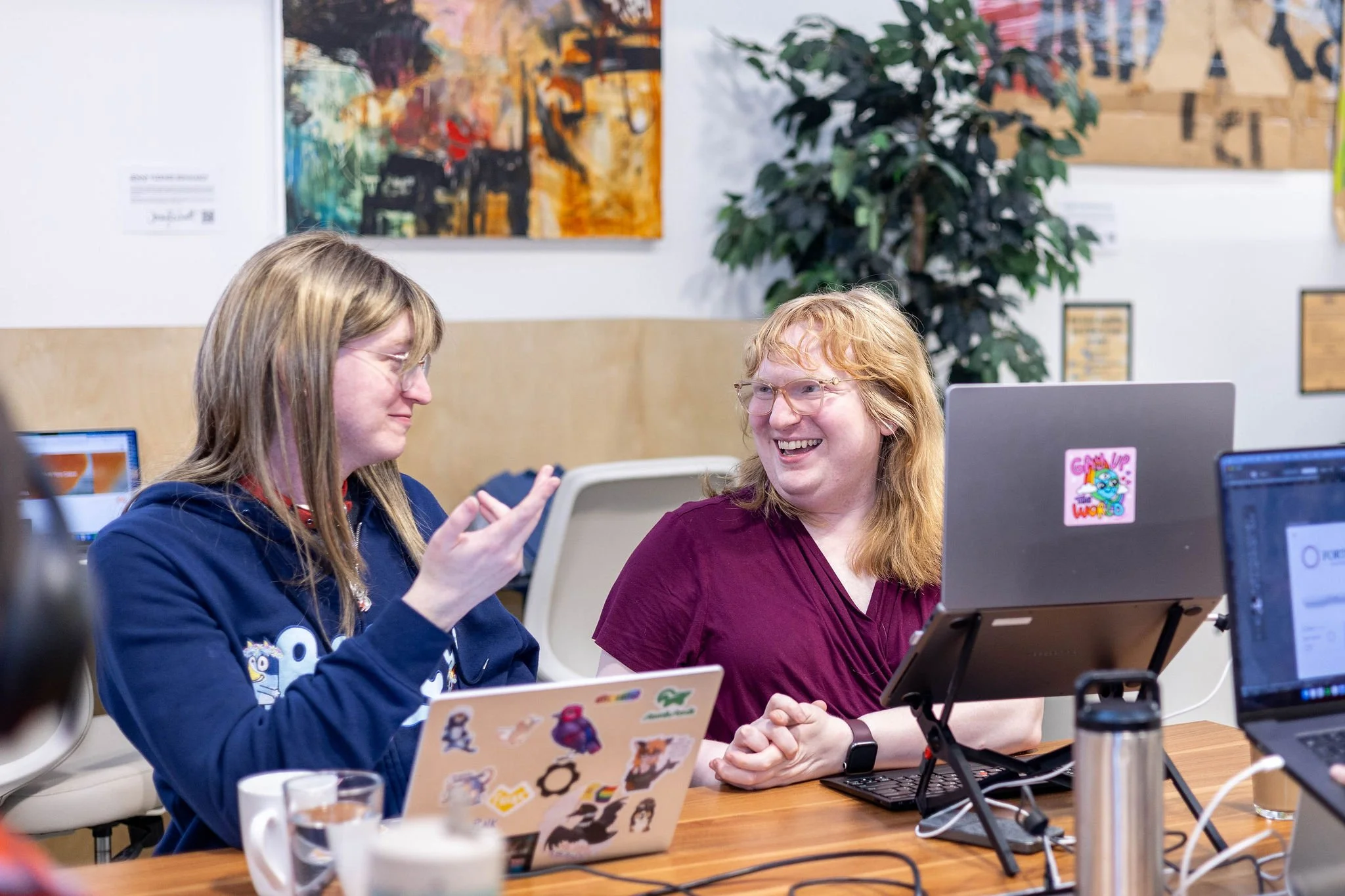 Two women smiling and talking in an office, one with glasses and the other with long hair, surrounded by laptops, one with stickers, and a large monitor, with abstract art and a plant in the background.