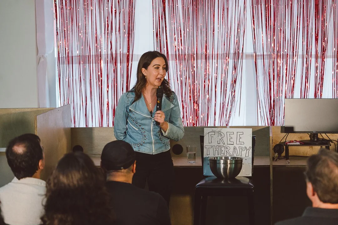 A woman in a blue jacket speaking into a microphone at a community event with a sign that says 'Free Therapy' and a metal bowl on a stool.