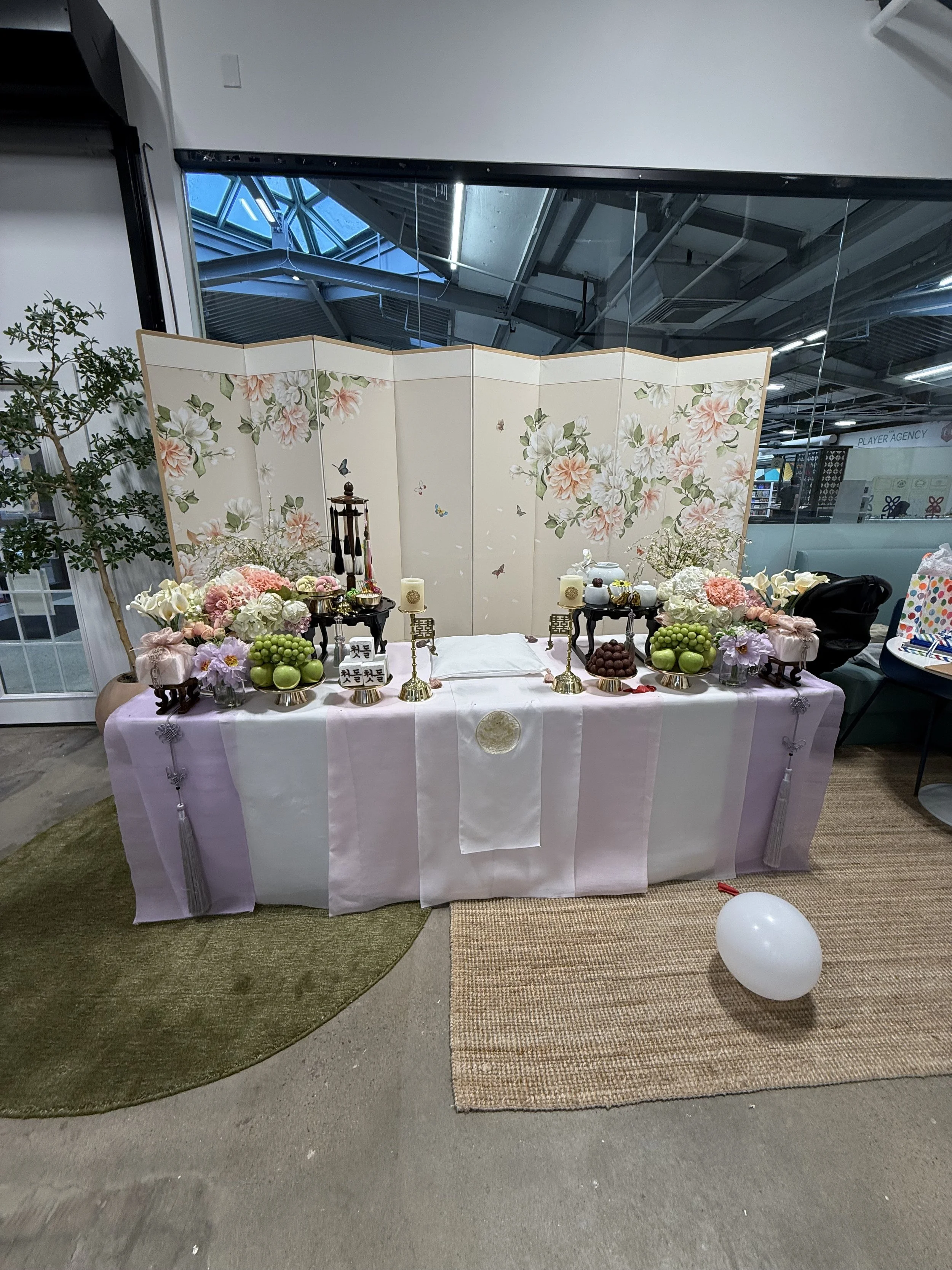 A decorative display table with flowers, fruits, and candles, set against a floral folding screen with butterflies, in an indoor space with a carpeted and concrete floor.