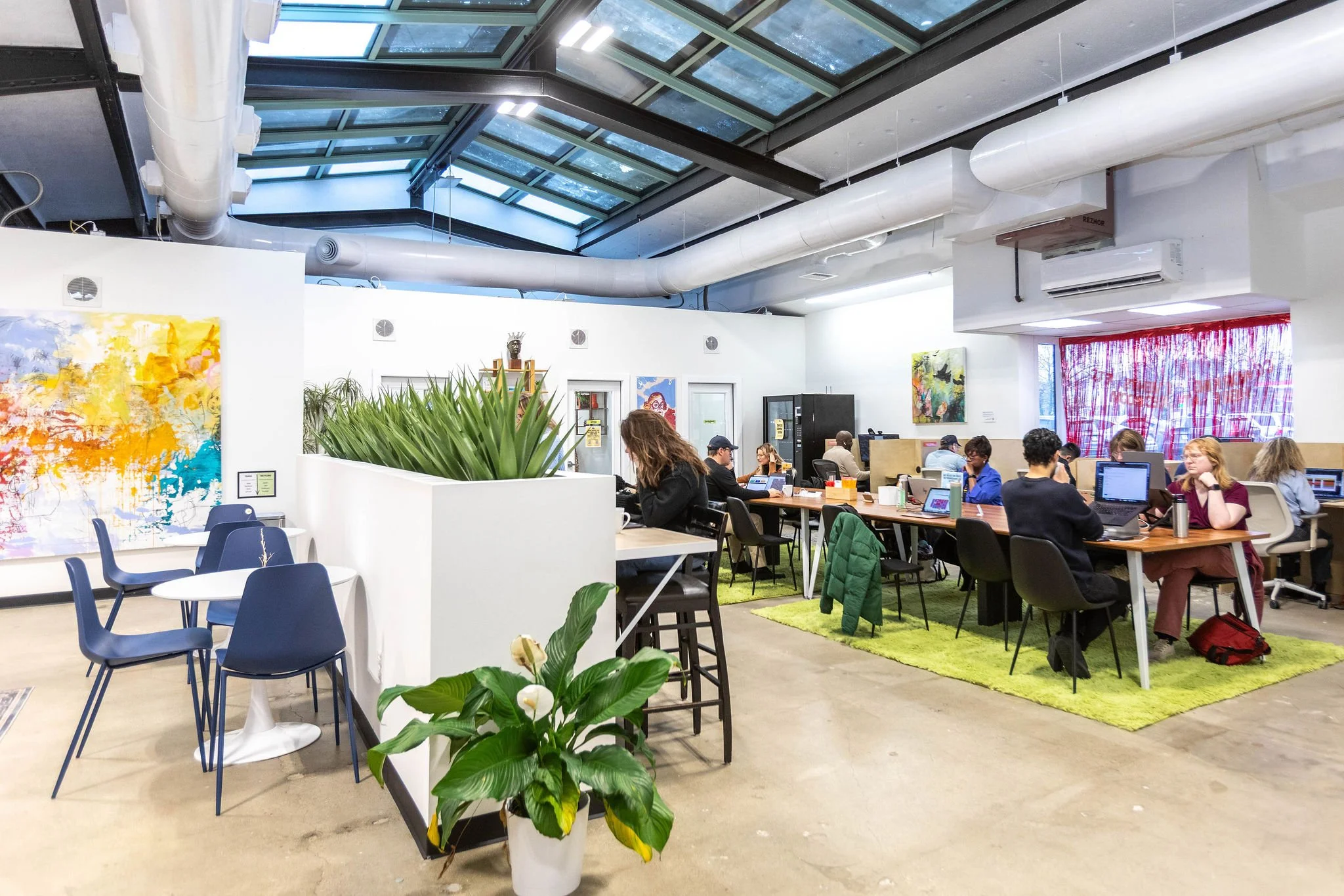 People working on laptops at a coworking space with modern decor, colorful artwork, green plants, and a large glass ceiling.