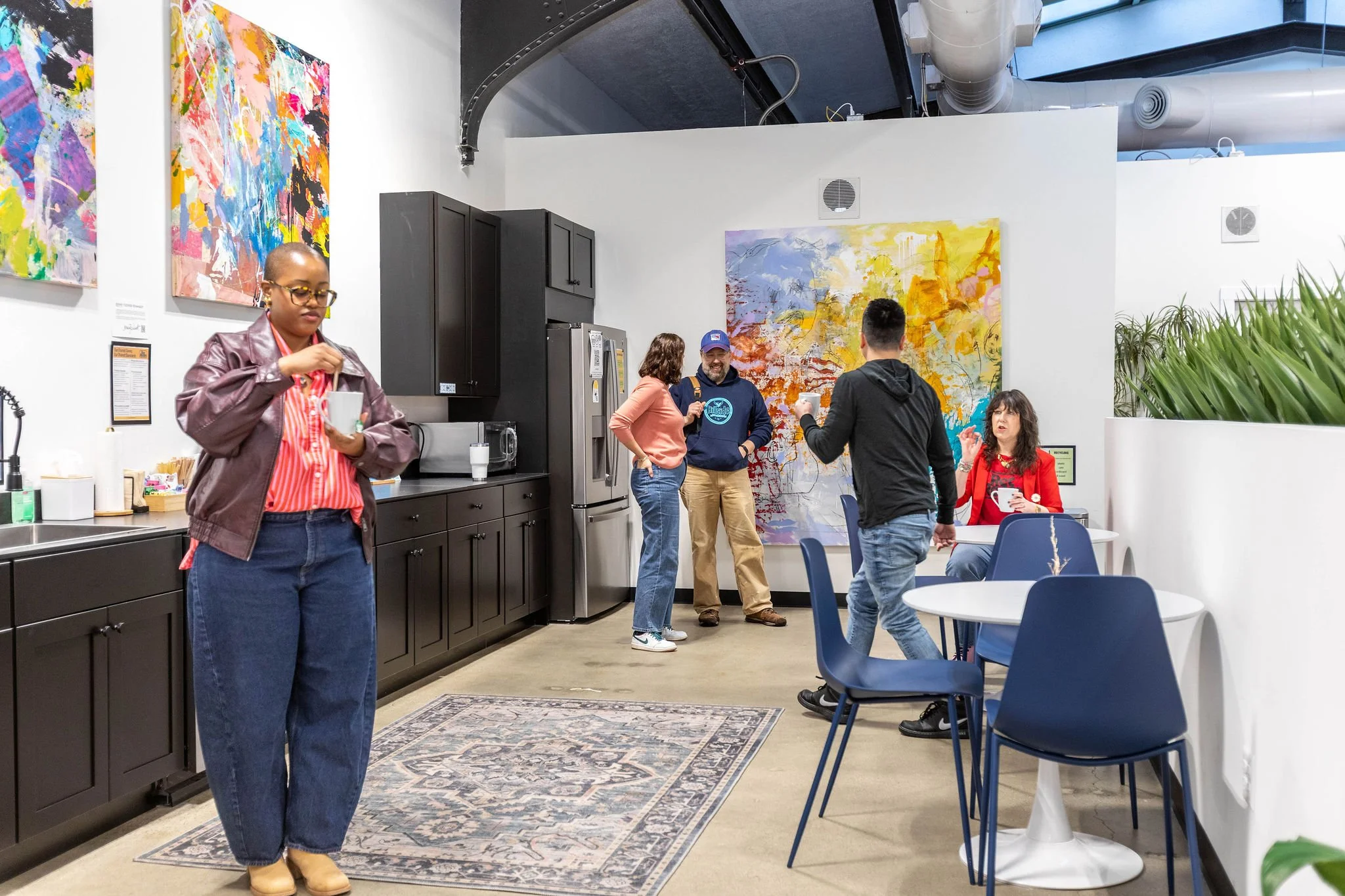 People socializing in a modern art gallery or cafe space, with colorful abstract paintings on the walls, black kitchen cabinets, and a mix of seating areas. A woman in the foreground is drinking from a mug, while others are engaged in conversation an