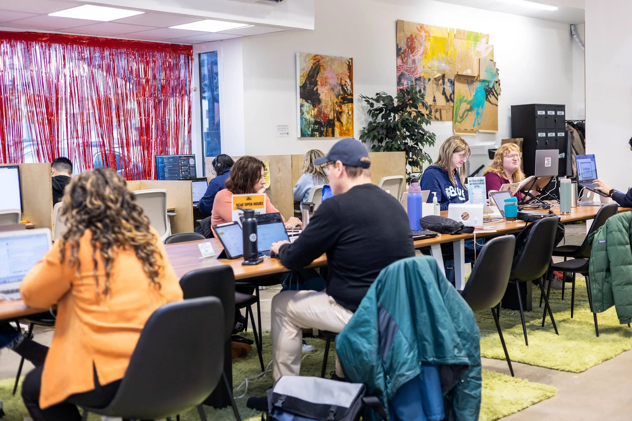 People working at a shared workspace with computers, artwork on the walls, and a colorful red curtain.