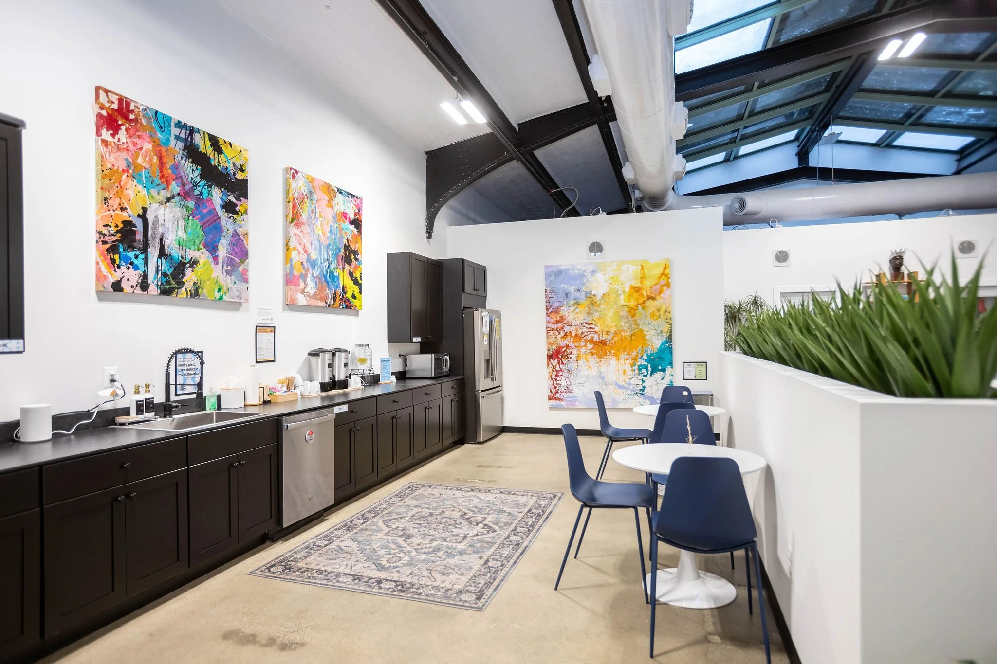 Interior of an art gallery or café with abstract paintings on the white walls, a black counter with coffee-making items, a refrigerator, small tables with blue chairs, a decorative rug on the floor, and a skylight ceiling.