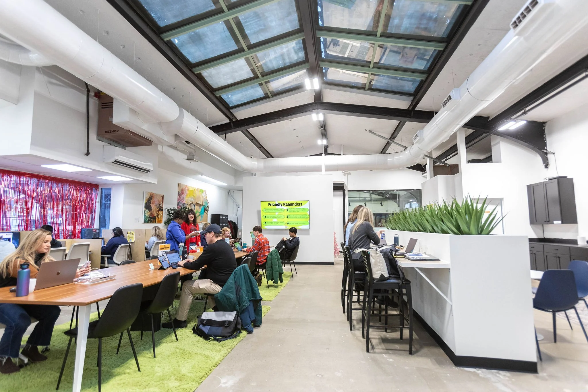 People working on laptops in a modern, open workspace with a glass ceiling, colorful wall art, and signs with reminders.