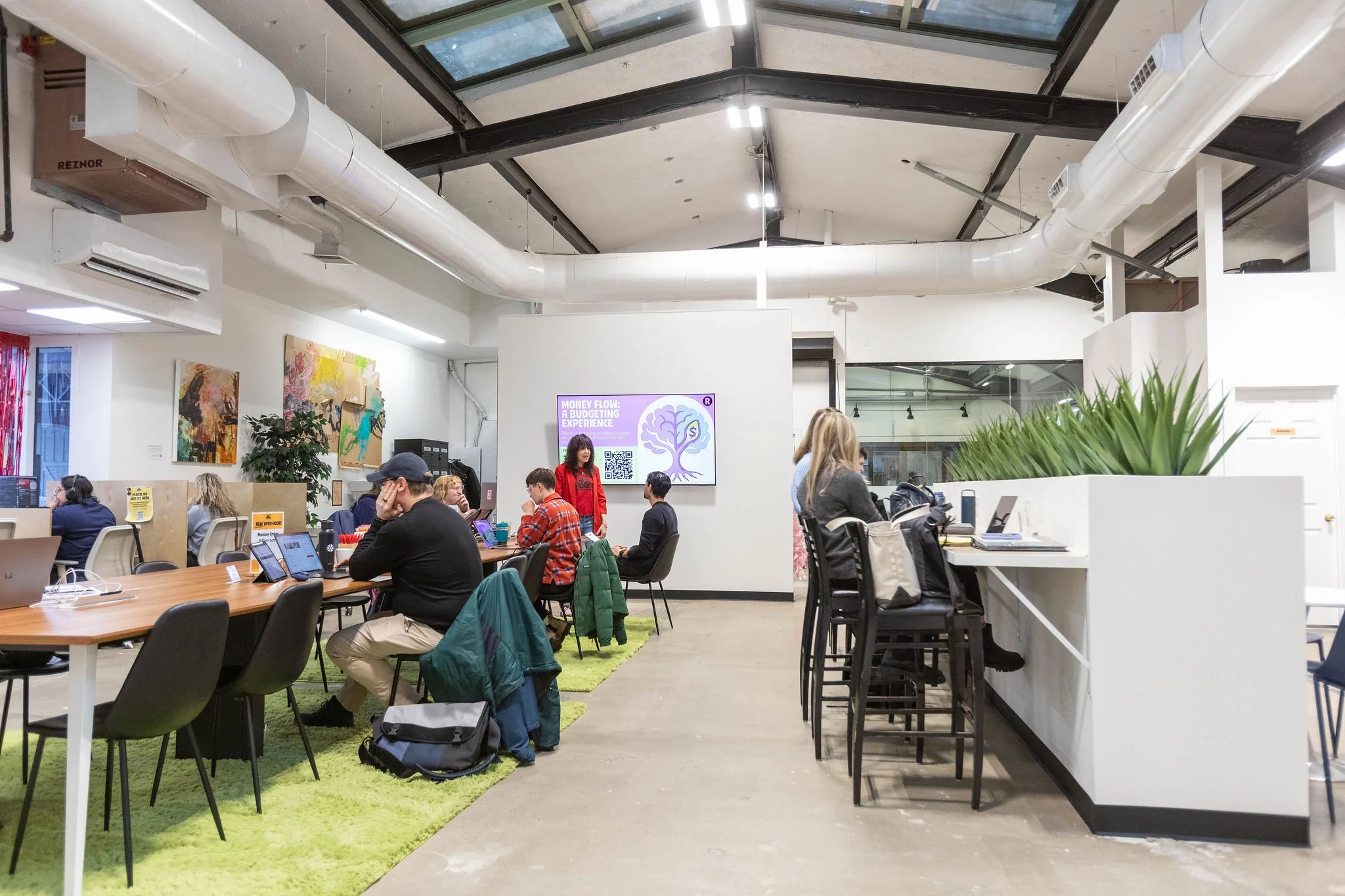 Open-concept coworking space with people working on laptops, a white wall with a digital screen, colorful abstract art, green plants, and a high ceiling with exposed ductwork.