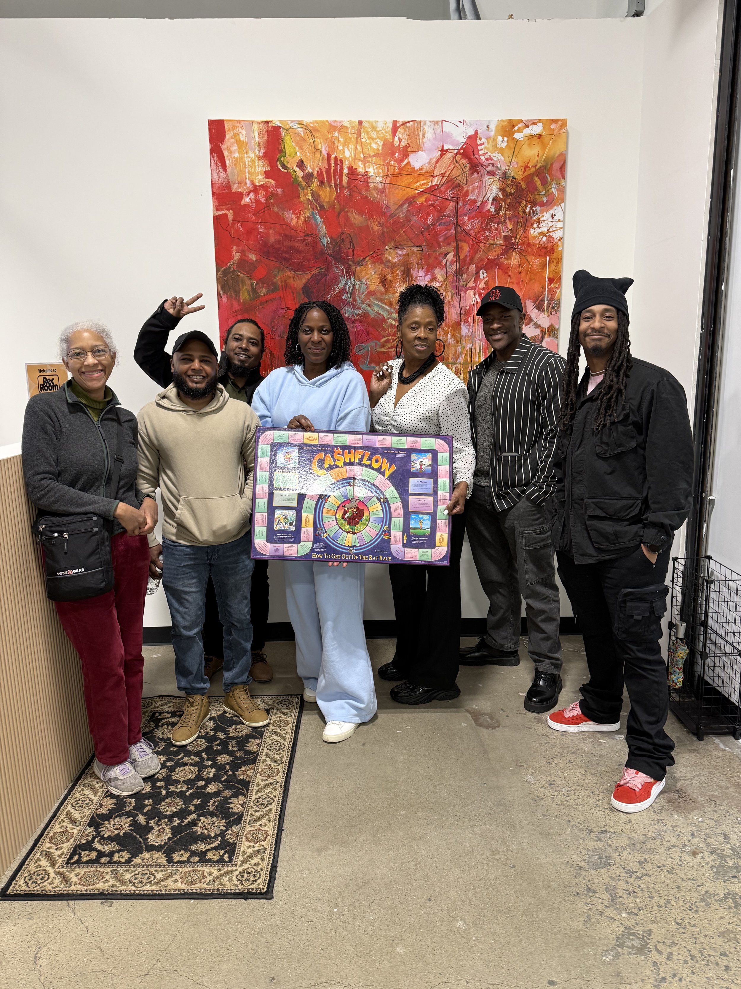Group of seven diverse people standing indoors in front of a colorful abstract painting, holding a board titled 'Cashflow' with game instructions, smiling and posing for the photo.