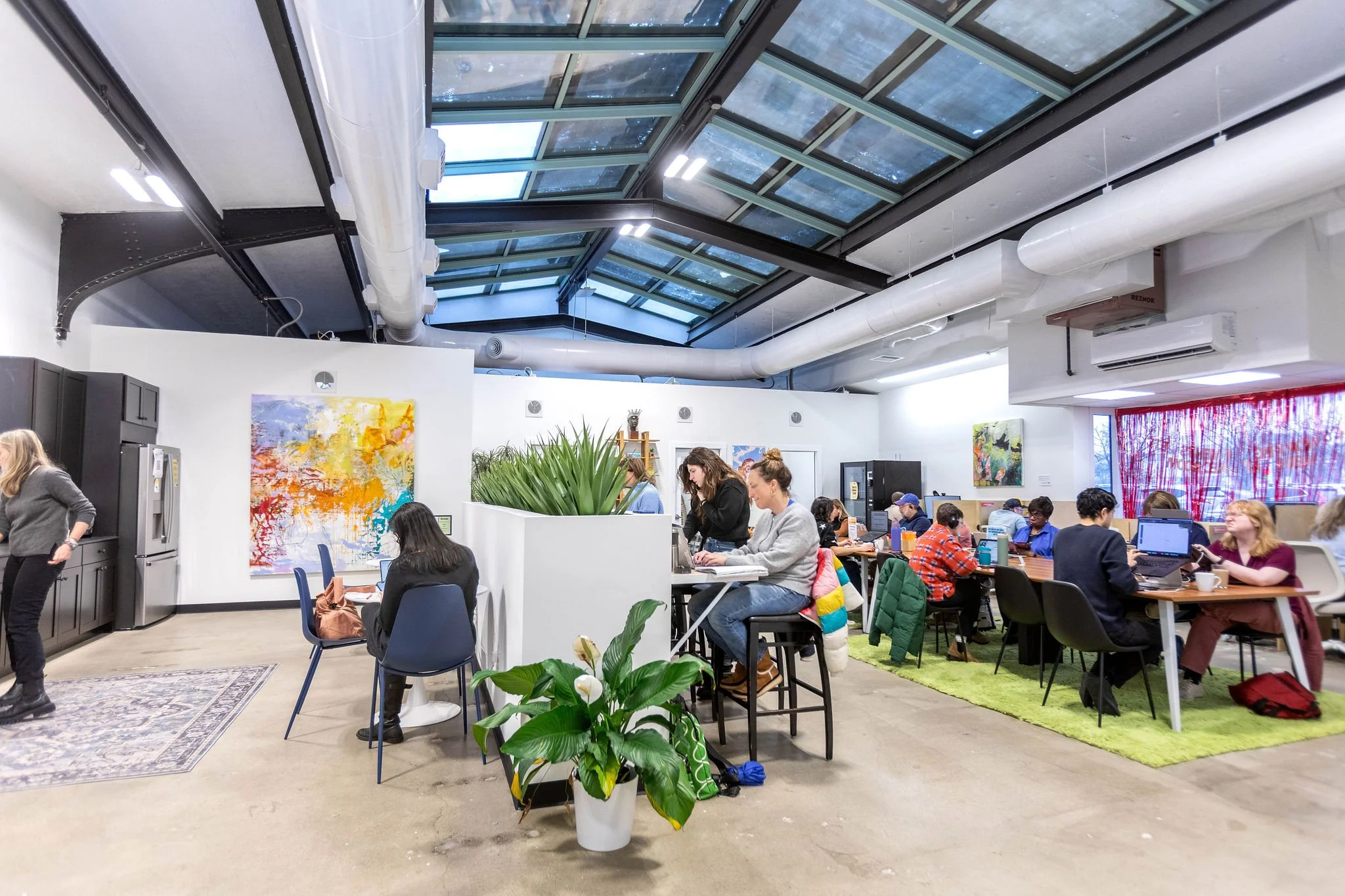 People working and socializing in a modern, open-coffee shop or coworking space with high glass ceiling, indoor plants, and colorful artwork on white walls.