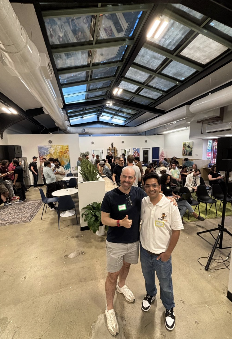 Two men standing together in a busy indoor event space, smiling at the camera. The man on the left is wearing a black t-shirt, beige shorts, and white sneakers, giving a thumbs-up. The man on the right is wearing a white polo shirt with a logo, blue jeans, and black and white sneakers. Behind them, people are seated and standing, engaging in conversations. The room has artwork on the walls, plants, and a glass ceiling with visible skylights.