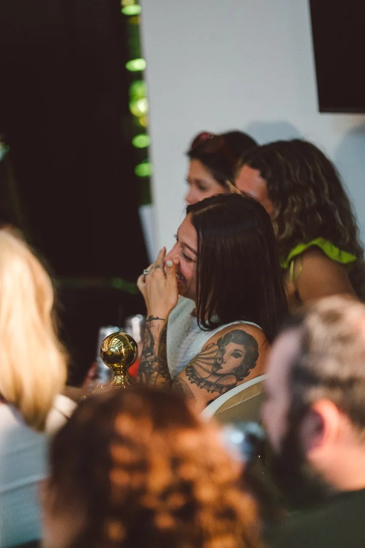 A woman with dark hair and tattoos on her arms is laughing and covering her mouth, sitting in a crowd at what appears to be an award show or event, with a golden trophy in front of her.