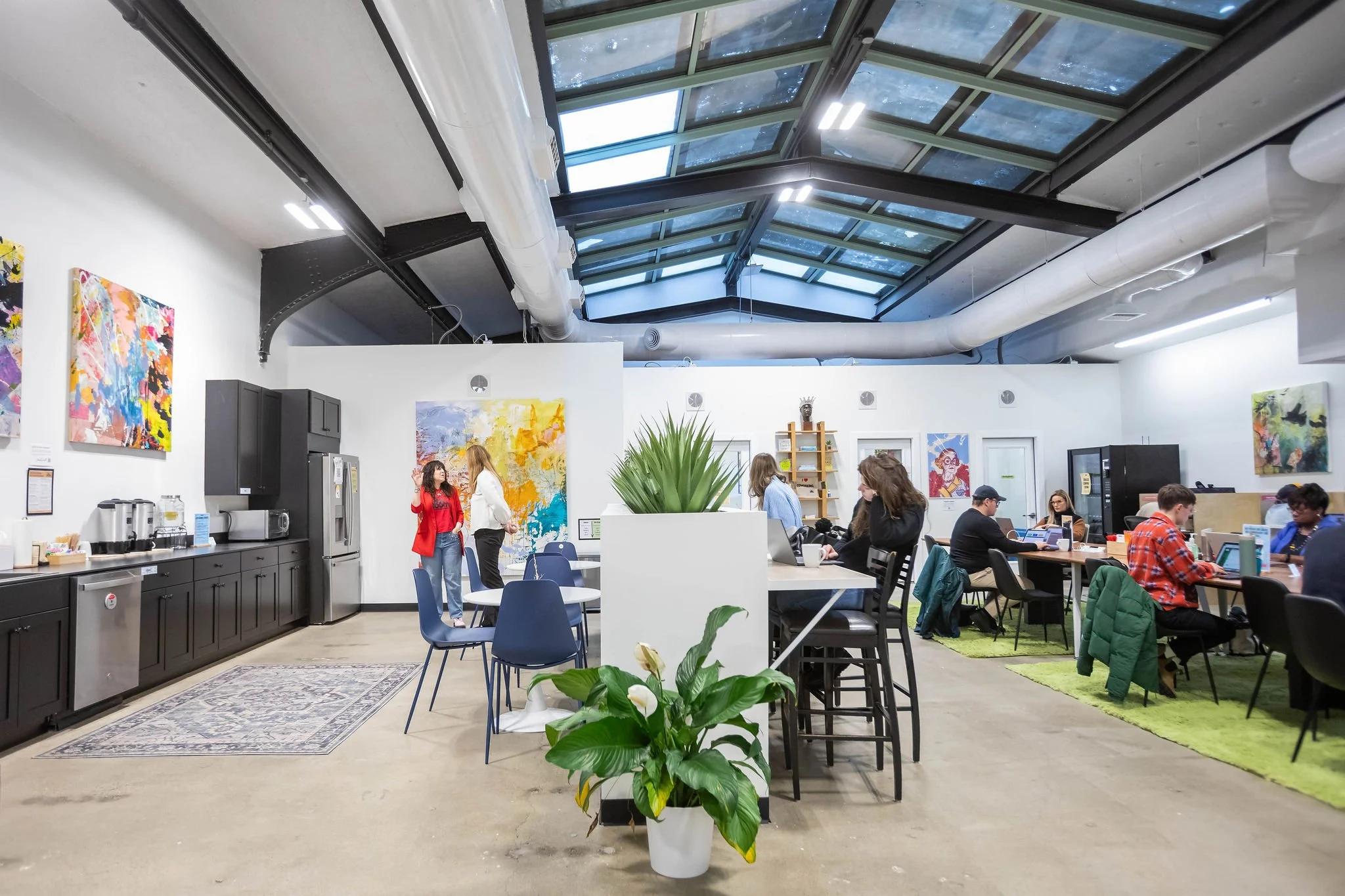 Modern coworking space with people working on laptops and socializing. Bright interior with colorful abstract art on white walls, large plant in the foreground, and a glass ceiling allowing natural light.