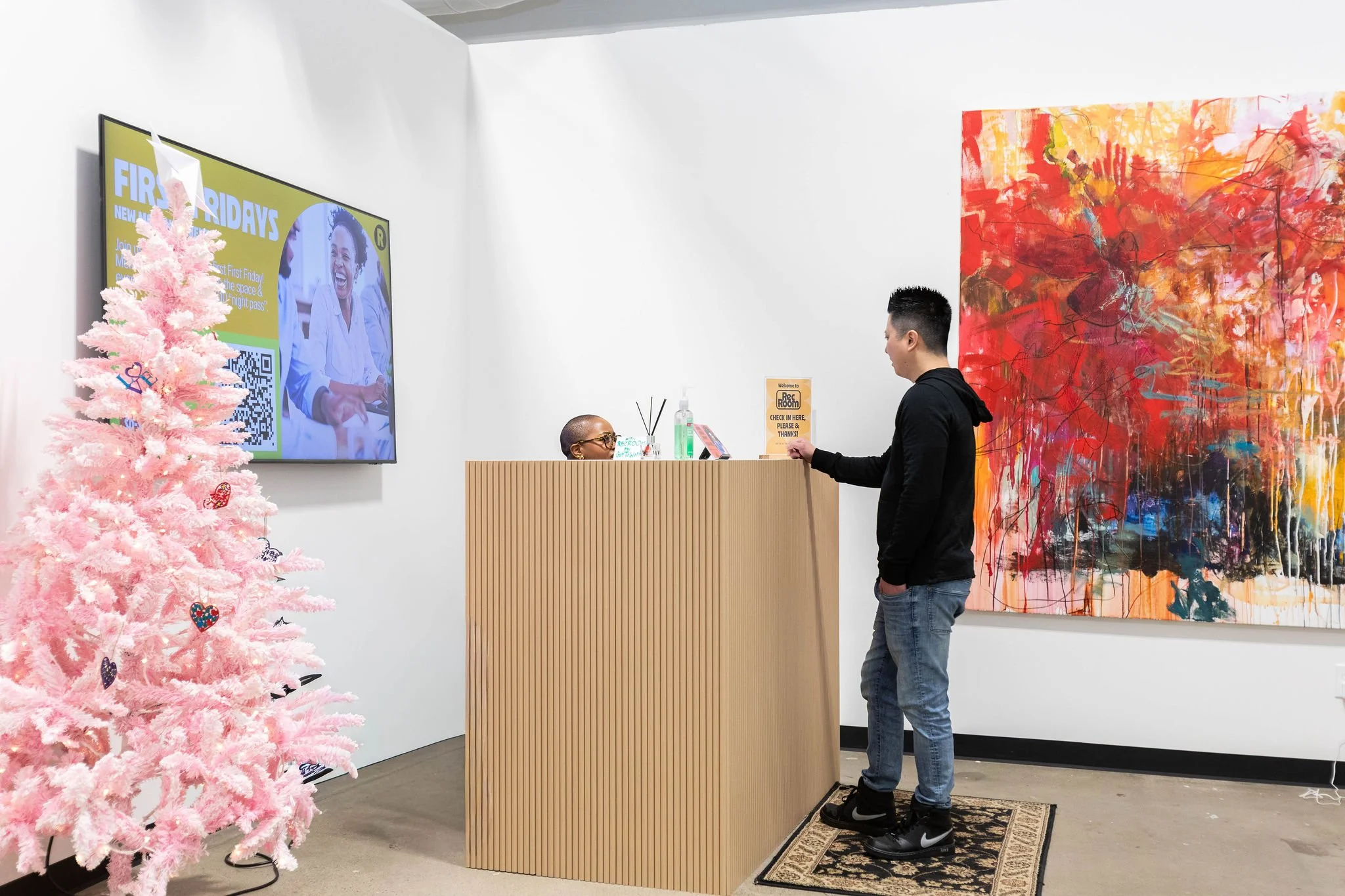 A man is checking in at a reception desk in an art gallery or office with a pink Christmas tree decorated with ornaments on the left, a large colorful abstract painting on the right, and a television screen displaying an advertisement on the wall behind the desk.