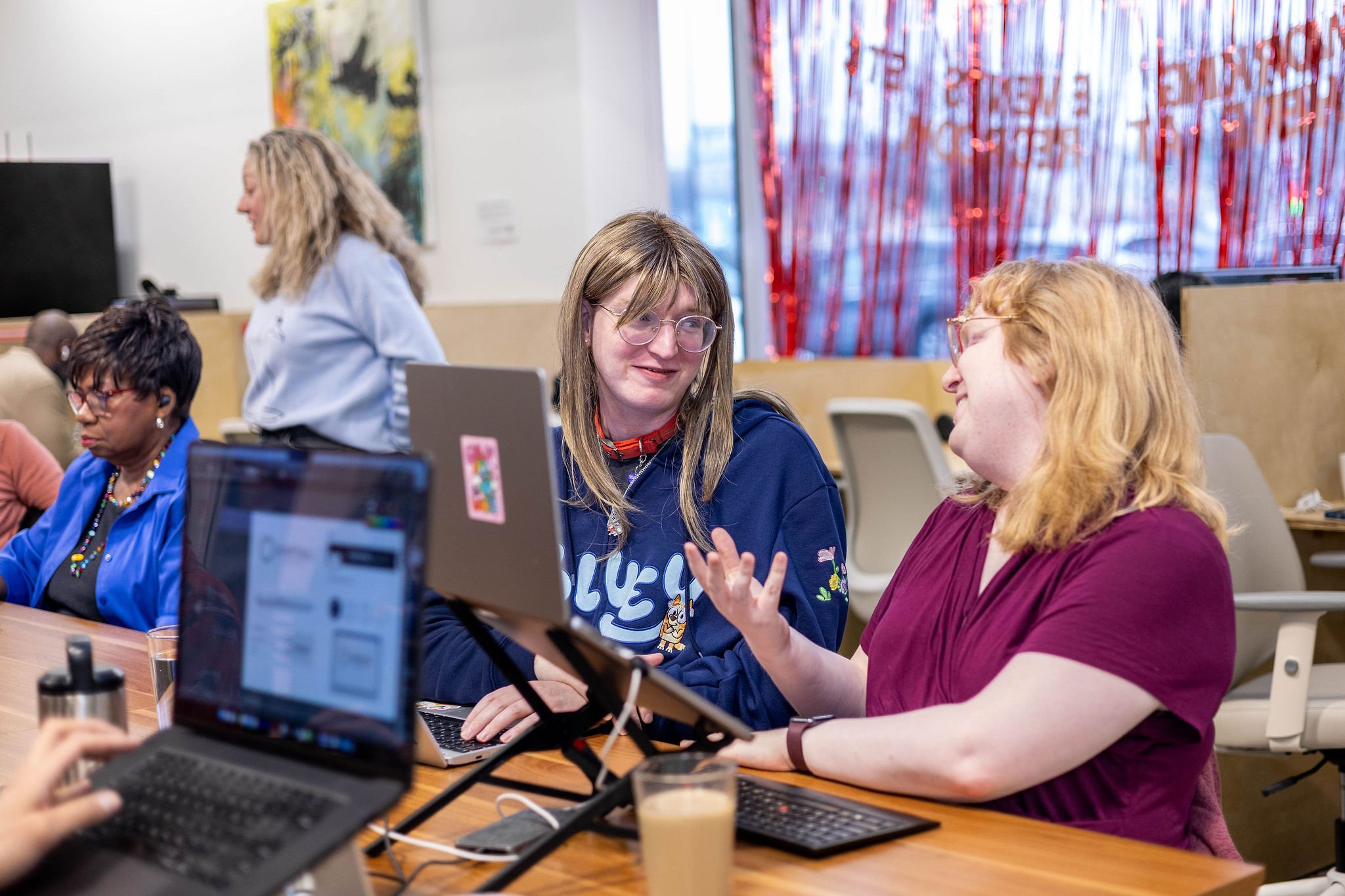 Two women sitting at a table engaged in conversation, with laptops and drinks in front of them, in a bright room with other people in the background.