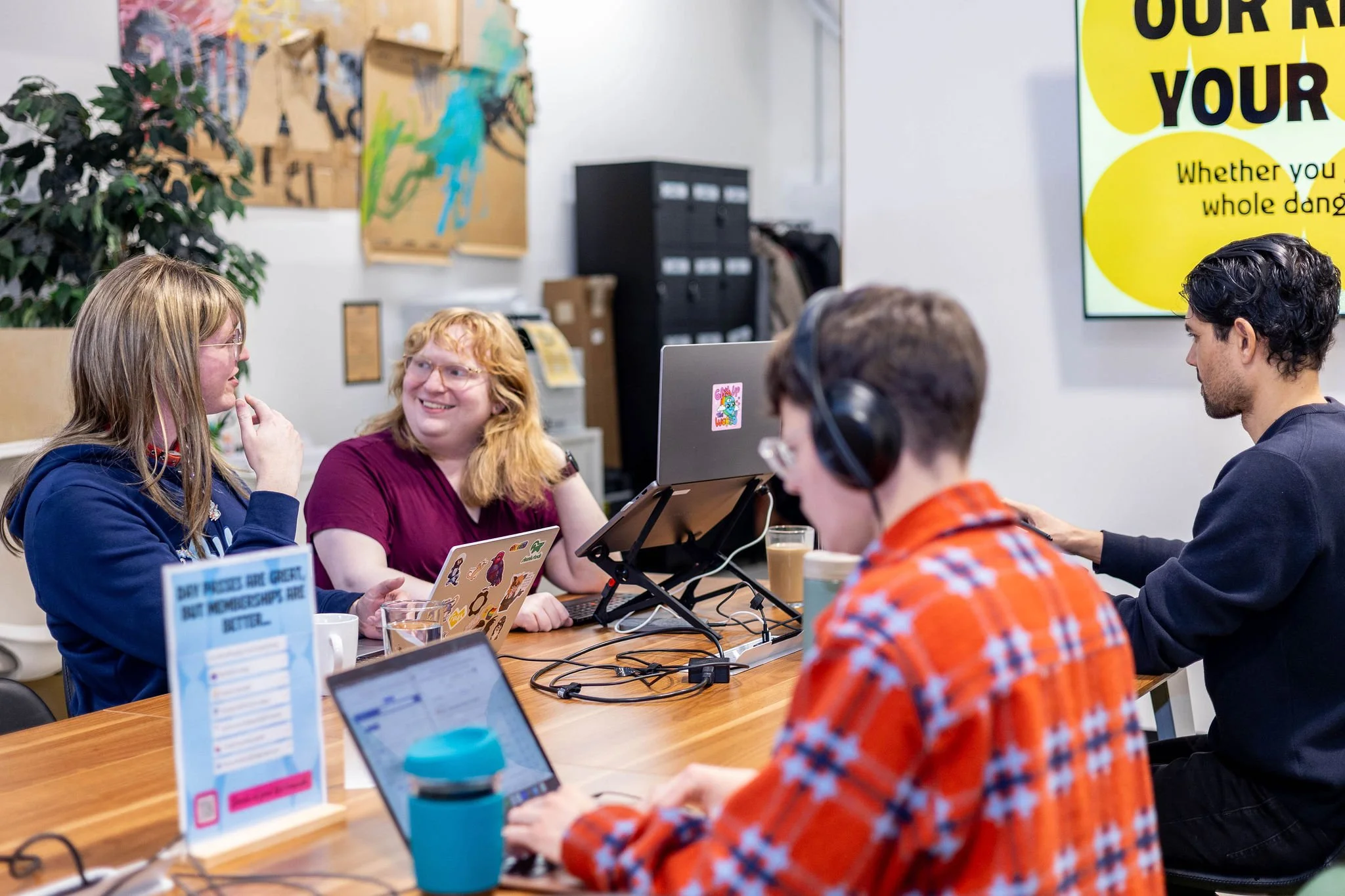 A group of five young adults working together at a wooden table in an office. They are using laptops and wearing headphones, and seem engaged in conversation. The background shows cardboard boxes, a large plant, artwork, and a bright yellow sign on a