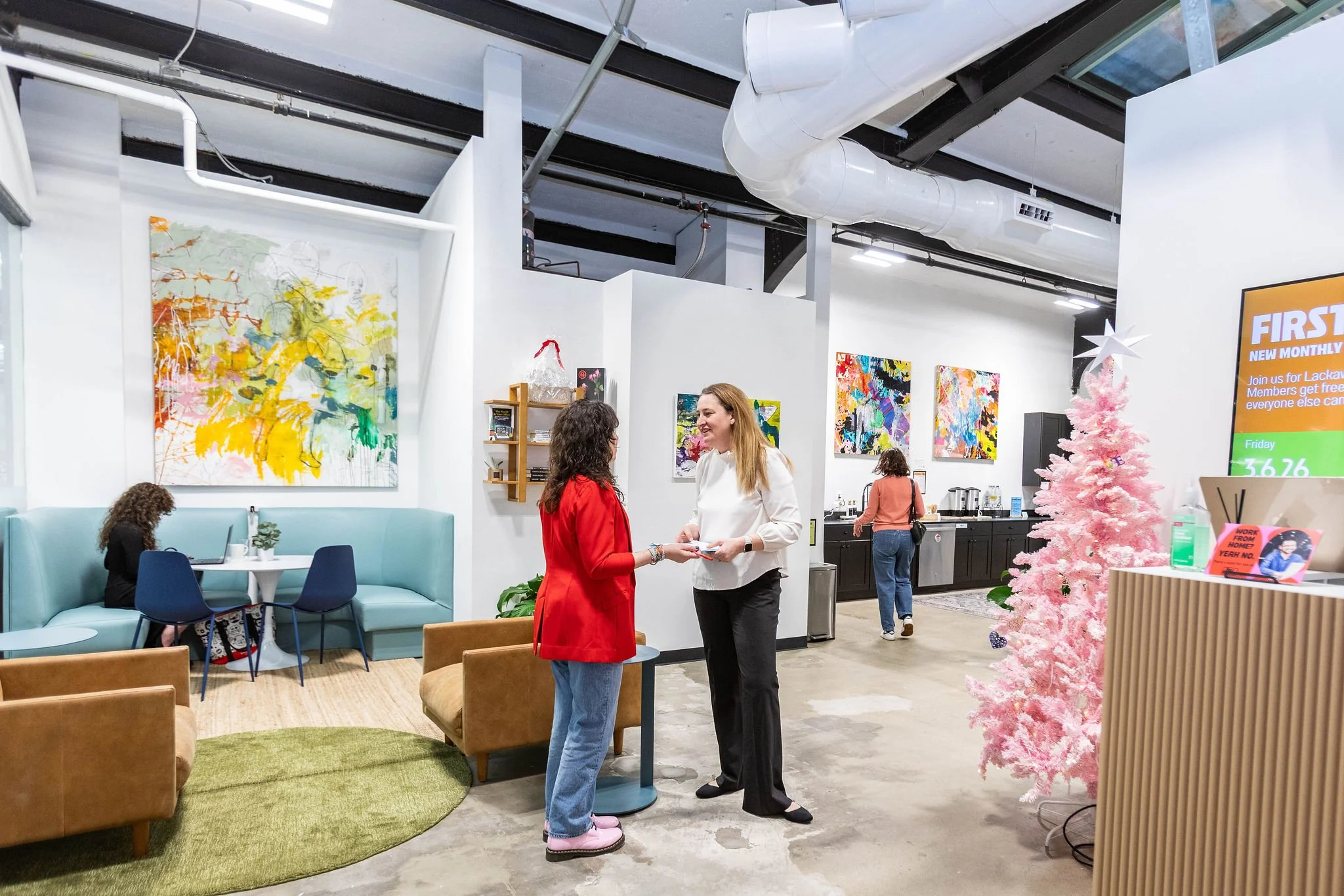 A brightly lit cafe or lounge area with abstract paintings on the walls, a pink Christmas tree, and people engaging in conversation and working on laptops.