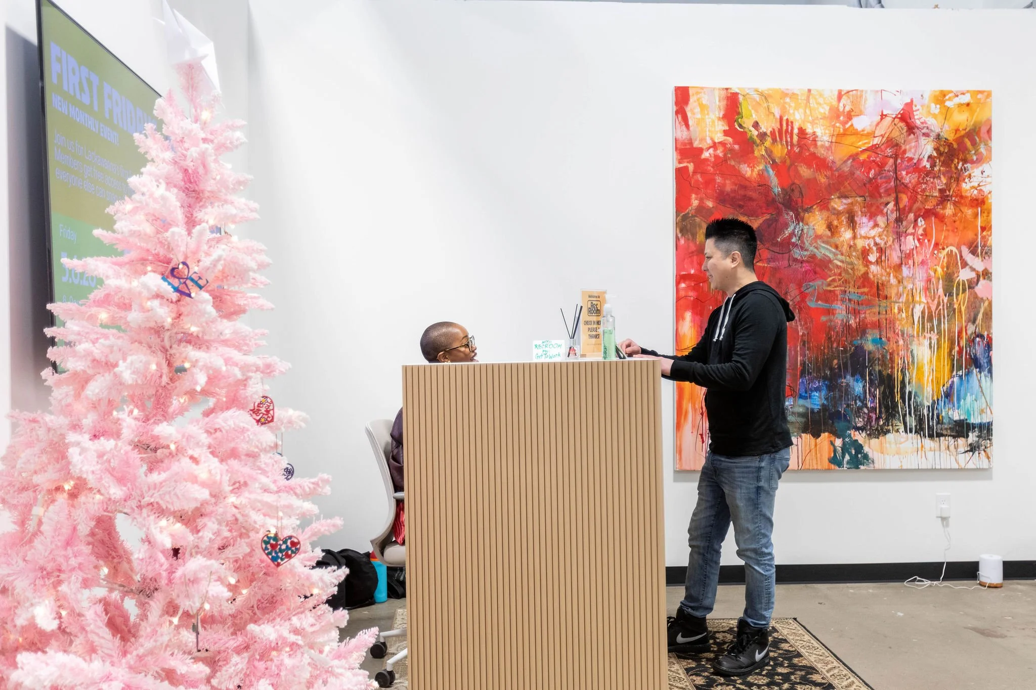 A man checks in with a woman sitting behind the desk. The room has a large, colorful abstract painting on the wall and a pink Christmas tree decorated with ornaments and ornaments. A large digital screen on the wall displays a promotional event.