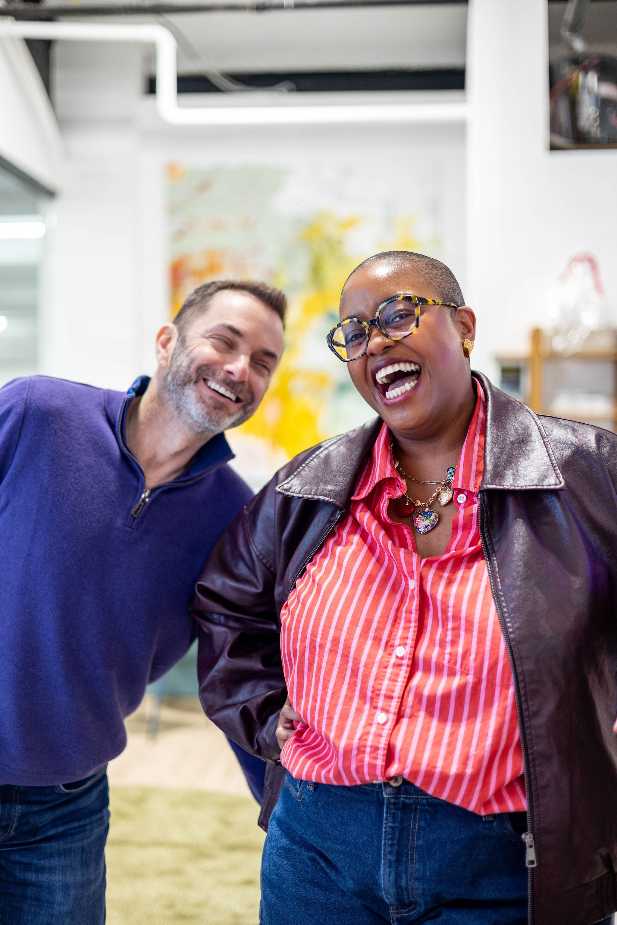Two people smiling and laughing indoors, a man with a beard wearing a blue sweater, and a woman wearing glasses, a leather jacket, and a red striped shirt.