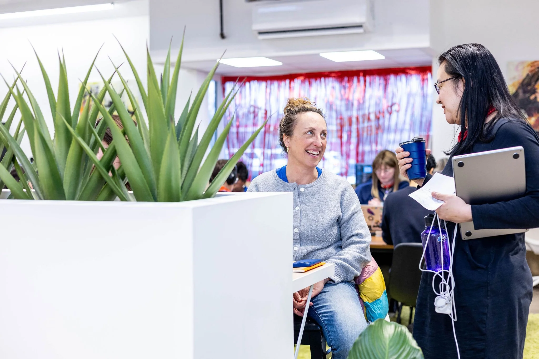 Two women are talking in a crowded indoor space, with one sitting and the other standing. The woman sitting has curly hair tied up and is smiling, while the woman standing has long black hair, glasses, and is holding a coffee tumbler, papers, and a l