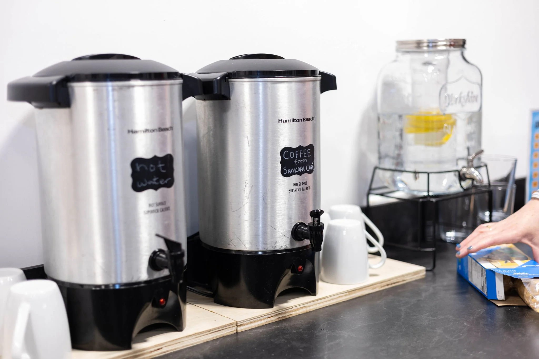Two stainless steel coffee dispensers labeled 'hot water' and 'coffee from Sankofa Cafe,' on a countertop with white mugs in front and a water dispenser to the right.