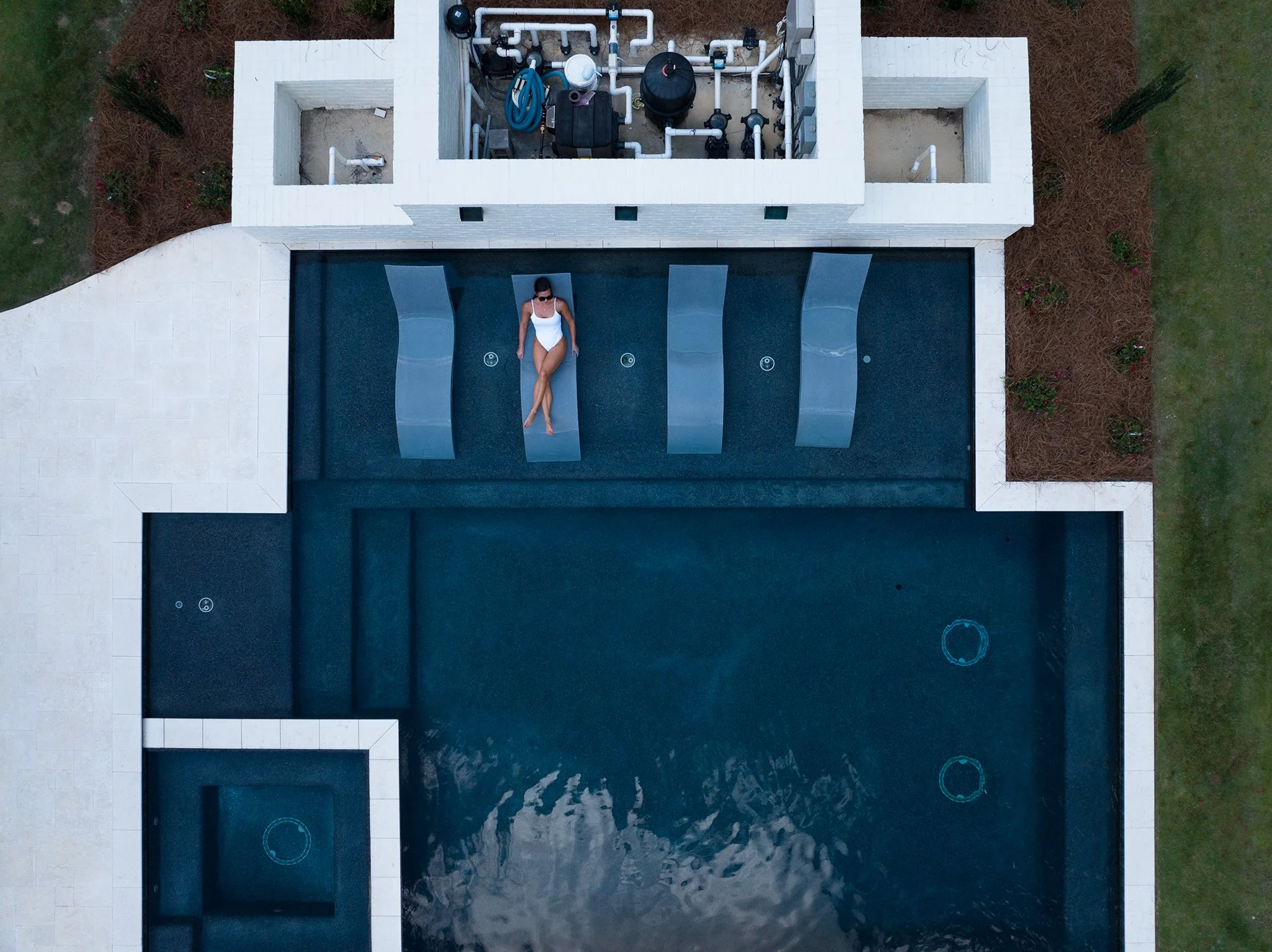 A woman in a white swimsuit and sunglasses is lounging on a sunbed beside a swimming pool, viewed from above. The pool area is surrounded by greenery and a white building with an open structure containing equipment and pipes.