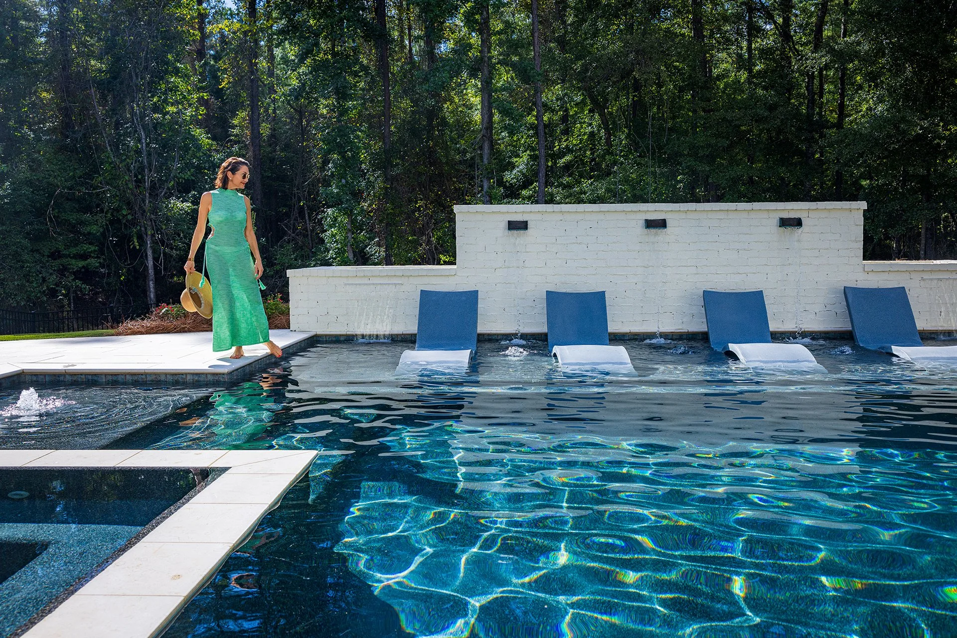 A woman in a green dress and sunglasses walking by a pool, holding a straw hat, with blue lounge chairs and a white brick wall with greenery behind.