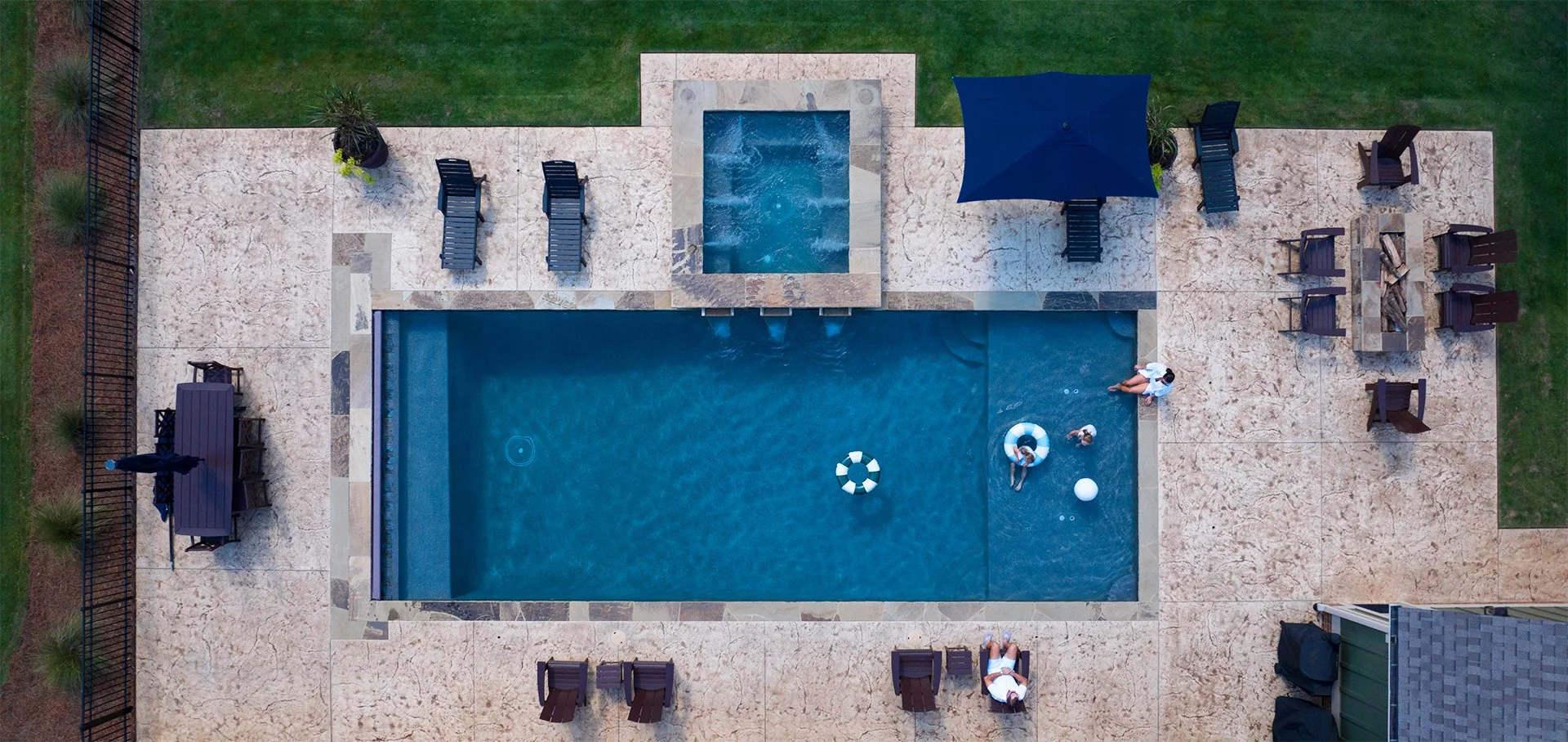 Aerial view of a backyard swimming pool area with several people, pool floats, lounge chairs, a table with chairs, an umbrella, and a hot tub, surrounded by grass and a fence.