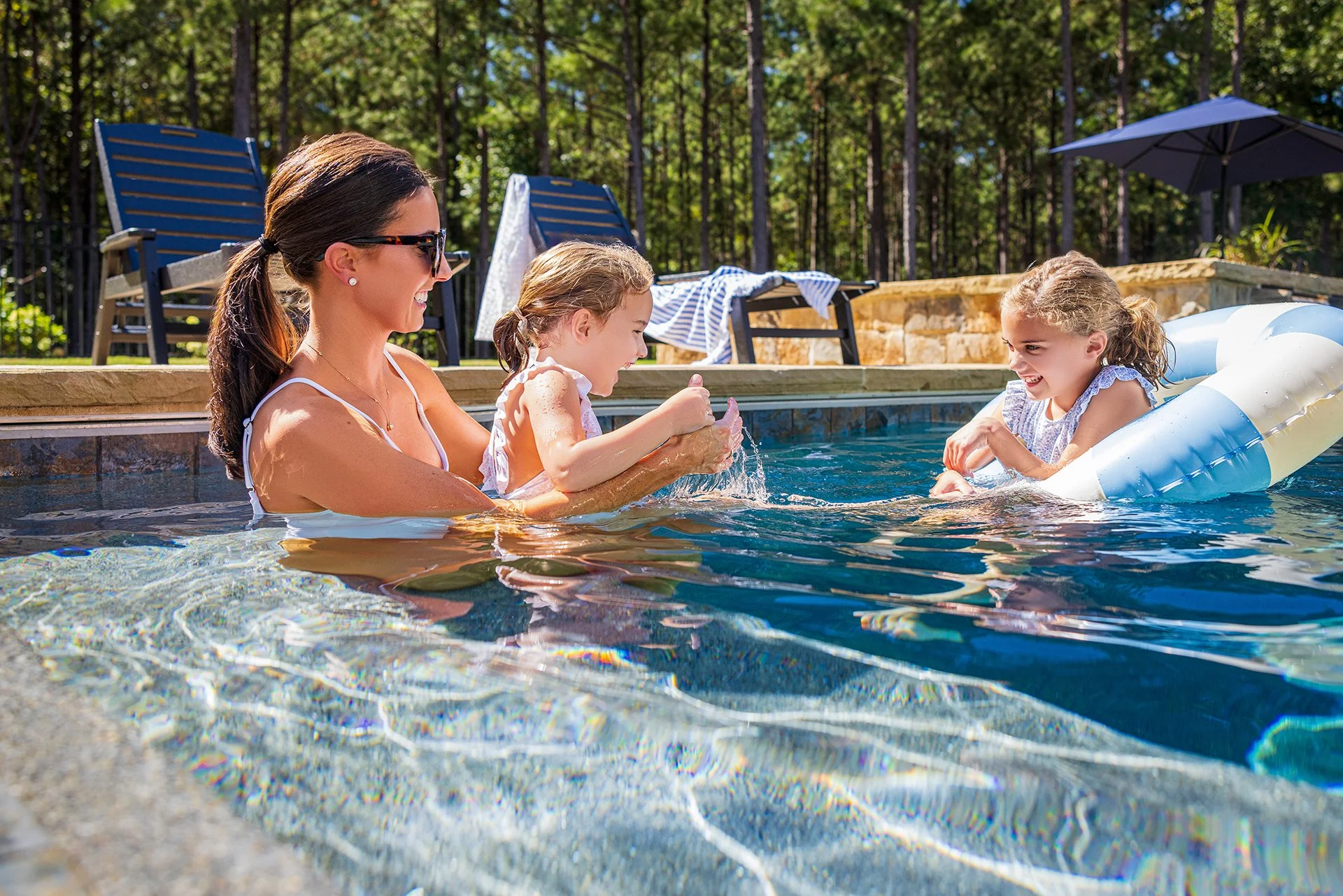 A woman and two young girls in a swimming pool enjoying a sunny day, with one girl playing and the other girl sitting on a float.