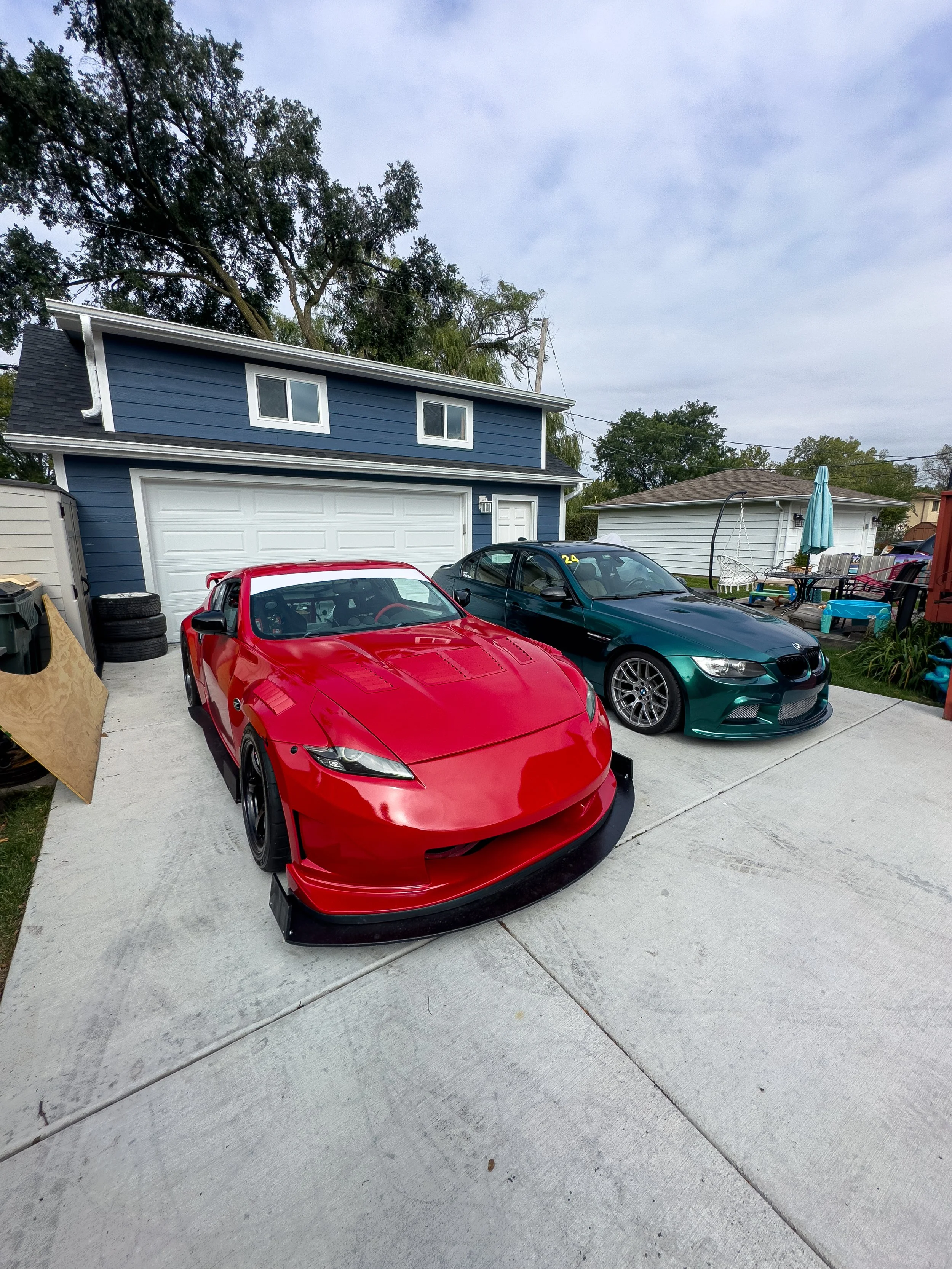 Two sports cars, a red one with a front splitter and a green one, parked in a driveway in front of a blue house with a garage. There are trees and outdoor furniture visible in the background.
