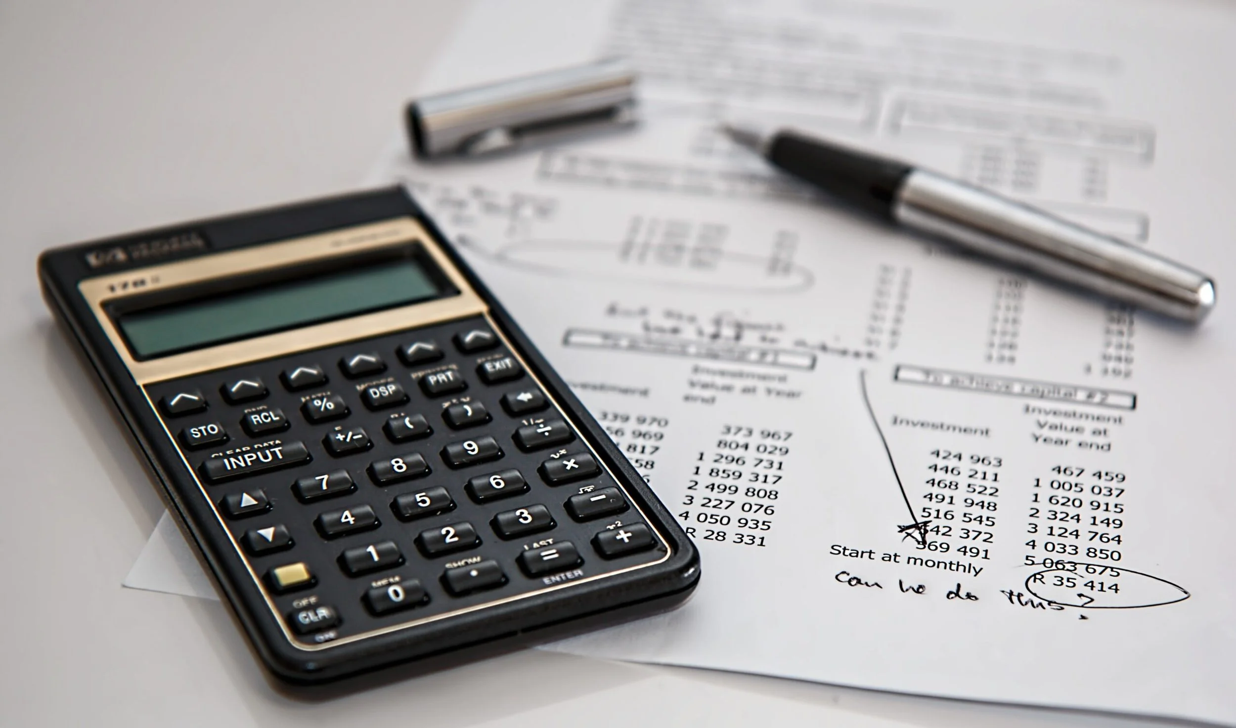 A calculator sits on top of a marked-up financial report that reads, 'Can we do this?' with a pen lying in the background.