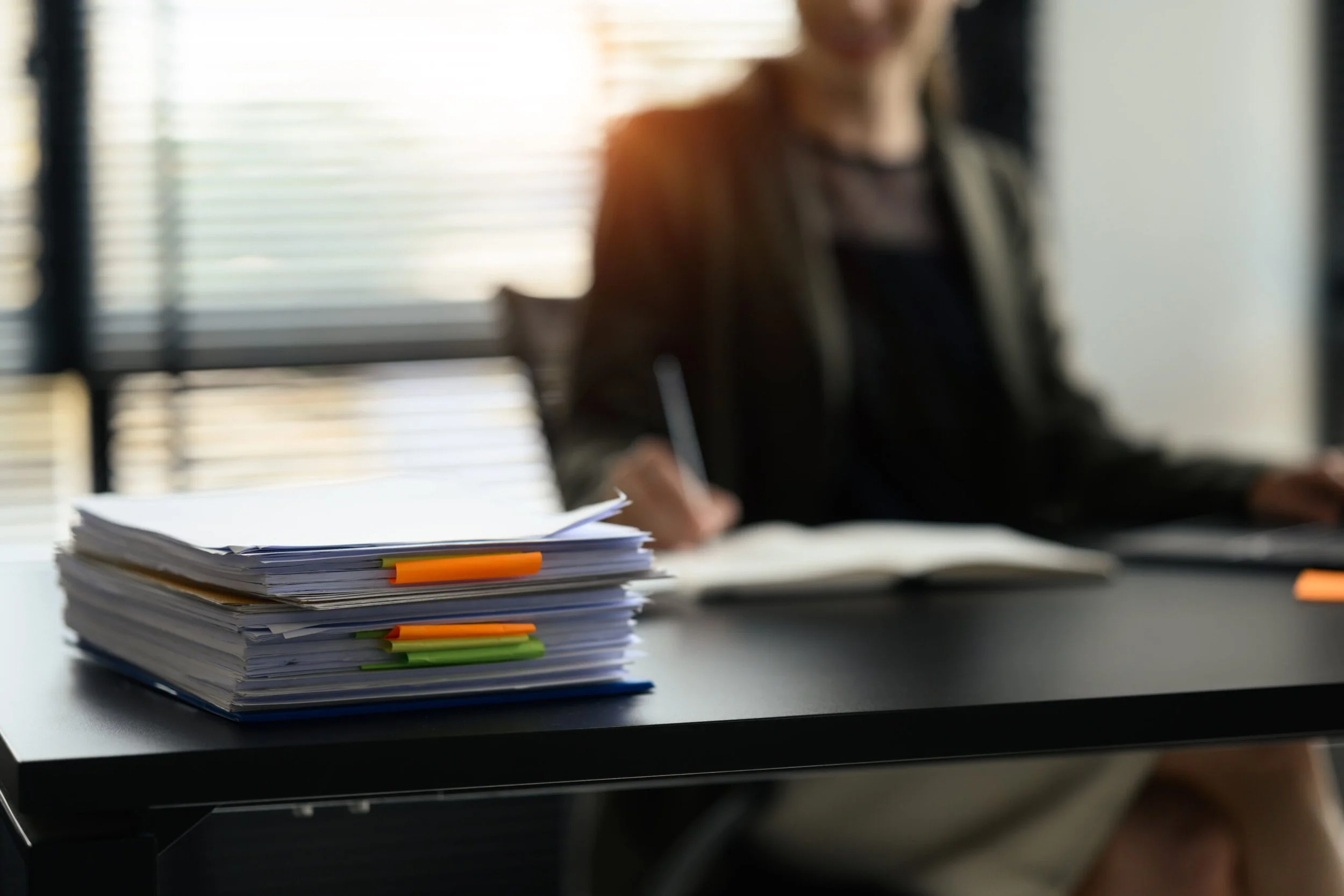 A person sitting at their desk, writing in a notebook next to a large stack of papers.