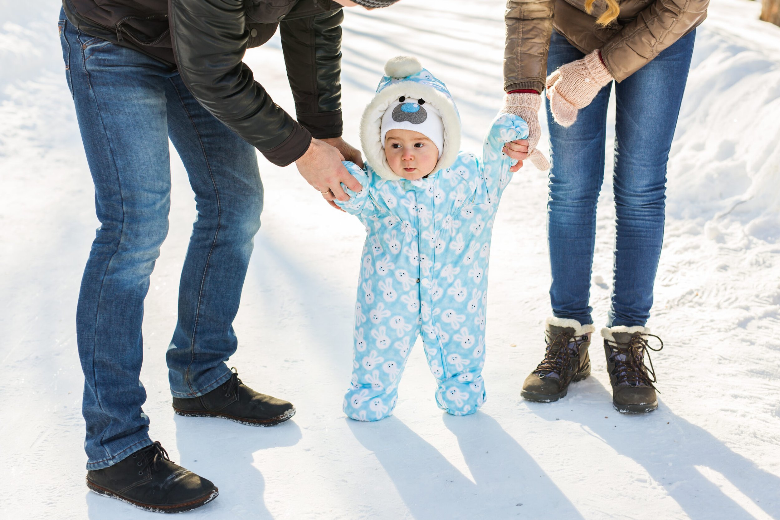 A baby in a snowsuit and hat taking steps while being supported by their mom and dad's hands.